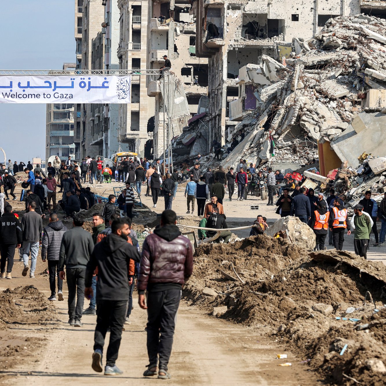 L'immagine mostra una scena in una zona colpita da conflitti, con edifici distrutti e rovine visibili sullo sfondo. Una grande insegna scritta in arabo e inglese dice "Benvenuti a Gaza". Diverse persone camminano lungo una strada, alcune indossano giubbotti arancioni, suggerendo che potrebbero essere soccorritori o volontari. L'atmosfera sembra essere quella di una comunità che cerca di ricostruirsi dopo una crisi.