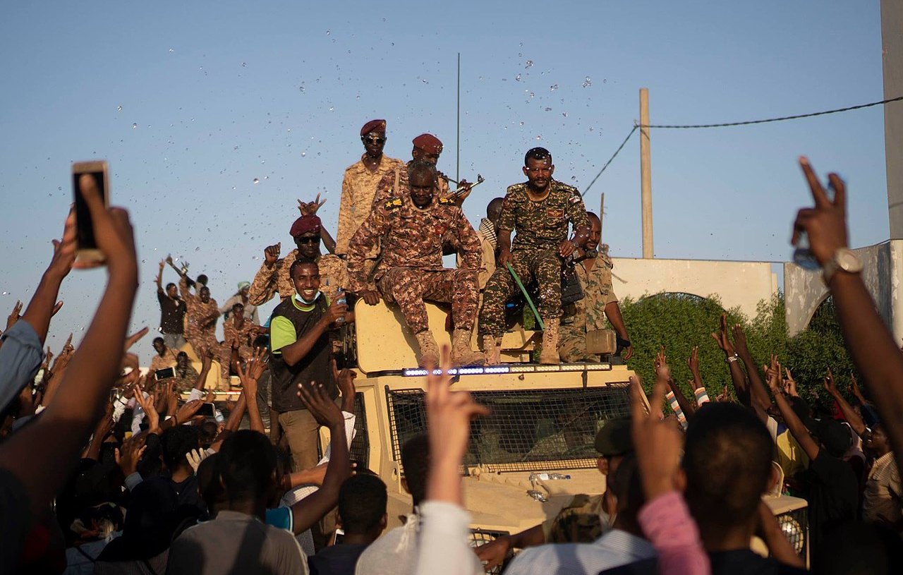 الصورة تظهر مجموعة من الجنود يقفون على متن مركبة عسكرية، بينما يبدو أن حشودًا من الناس تتجمع حولهم. يبدو الجو احتفاليًا حيث يرفع بعض الأشخاص أيديهم في الهواء، ويبدو أنهم يعبرون عن الفرح أو التأييد. السماء زرقاء وظهرت بعض الأعلام والاحتفالات في الخلفية.