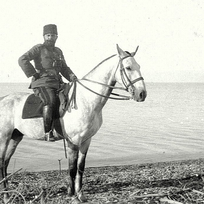 در تصویر، یک مرد uniform پوشیده بر روی یک اسب سفید ایستاده است. او در کنار آب قرار دارد و زمینه‌ای آرام و طبیعی دیده می‌شود. لباس او با جزئیات مشخصی همراه است و حالتی رسمی دارد. اسب نیز با ظاهر زیبا و شکیل خود توجه را جلب می‌کند. این صحنه به نظر می‌رسد در زمان گذشته و در مکانی نزدیک به دریا یا دریاچه ثبت شده باشد.