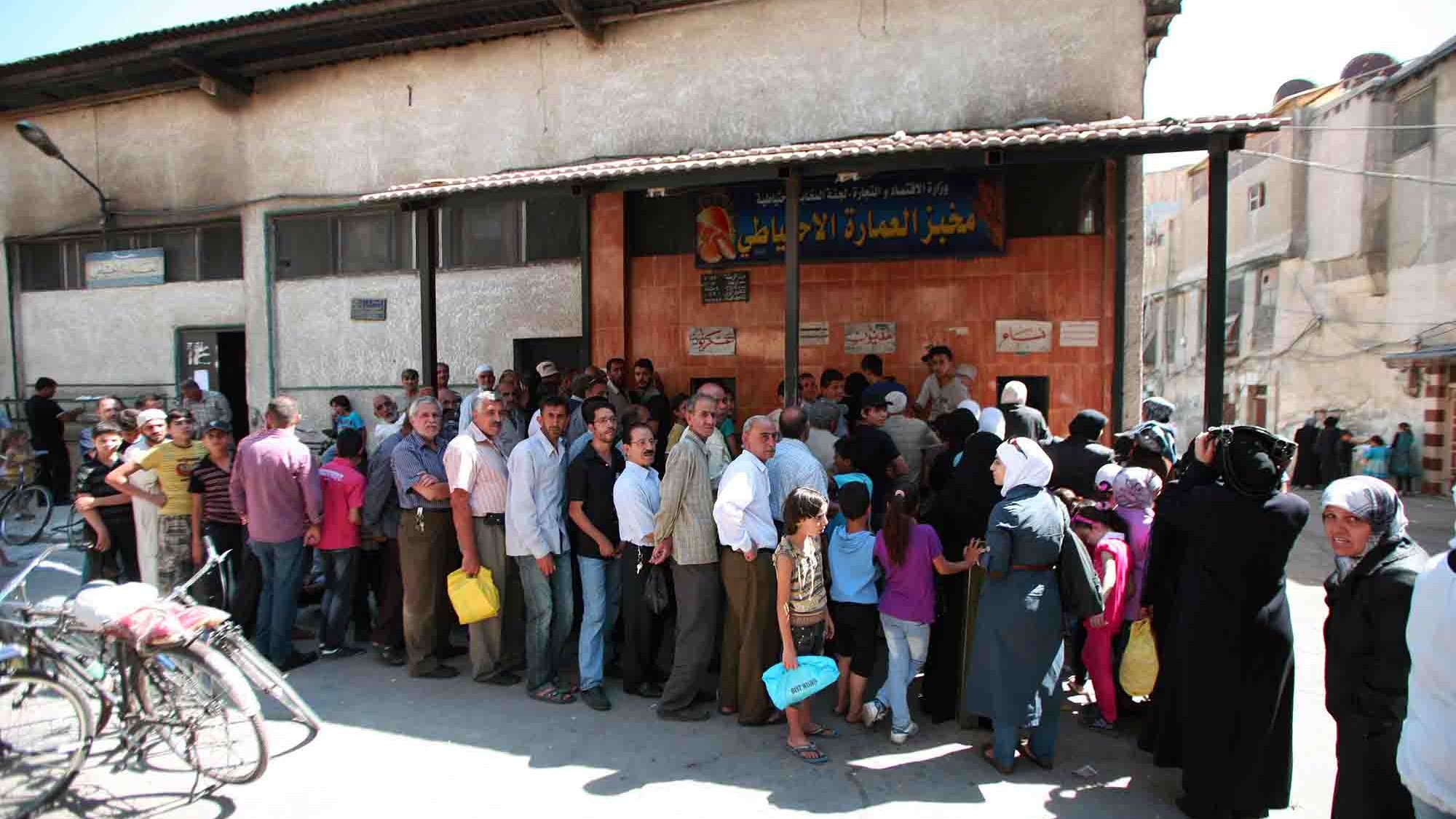 L'immagine mostra una lunga fila di persone che si trovano all'esterno di un edificio. La scena sembra essere in un contesto urbano, probabilmente in una città del Medio Oriente. Le persone nella fila sono di diverse età e indossano abiti vari, alcuni dei quali tradizionali. Alcuni individui portano sacche o borse, e si può notare un'atmosfera di attesa e di comunità. Sullo sfondo, ci sono edifici semplici con elementi architettonici tipici della regione.