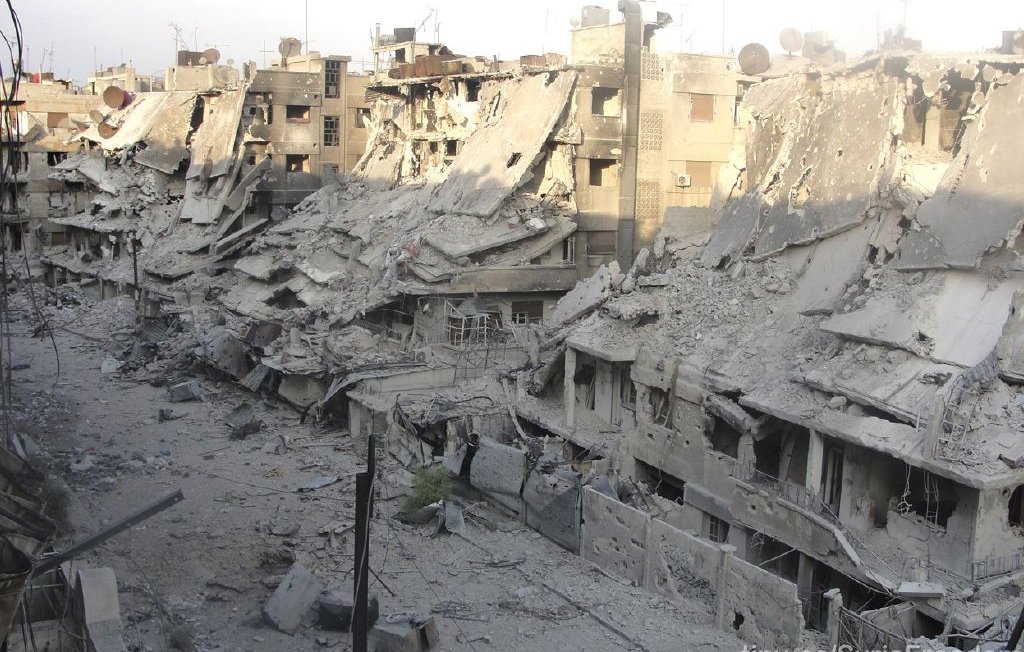 L'image montre un paysage urbain fortement endommagé, avec des bâtiments effondrés et en ruines. Les façades des immeubles sont détruites, laissant entrevoir des débris et des structures instables. Le sol est recouvert de débris et de poussière, témoignant de la violence des destructions. La lumière du jour éclaire la scène, accentuant le contraste entre les ruines et le ciel. Cette image évoque un environnement de guerre et de crise humanitaire.