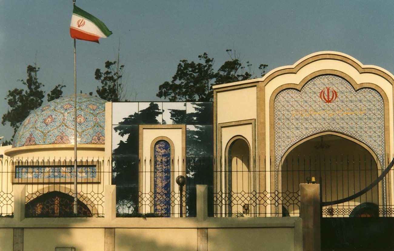 L'image montre un bâtiment architectural de style iranien, typique avec des tuiles colorées et des motifs ornementaux. On peut voir le drapeau iranien flottant sur un mât à gauche. Le bâtiment présente une structure avec un dôme décoré et des arcs, intégrant des éléments de design traditionnels. L'environnement est arboré, avec des arbres visibles en arrière-plan.