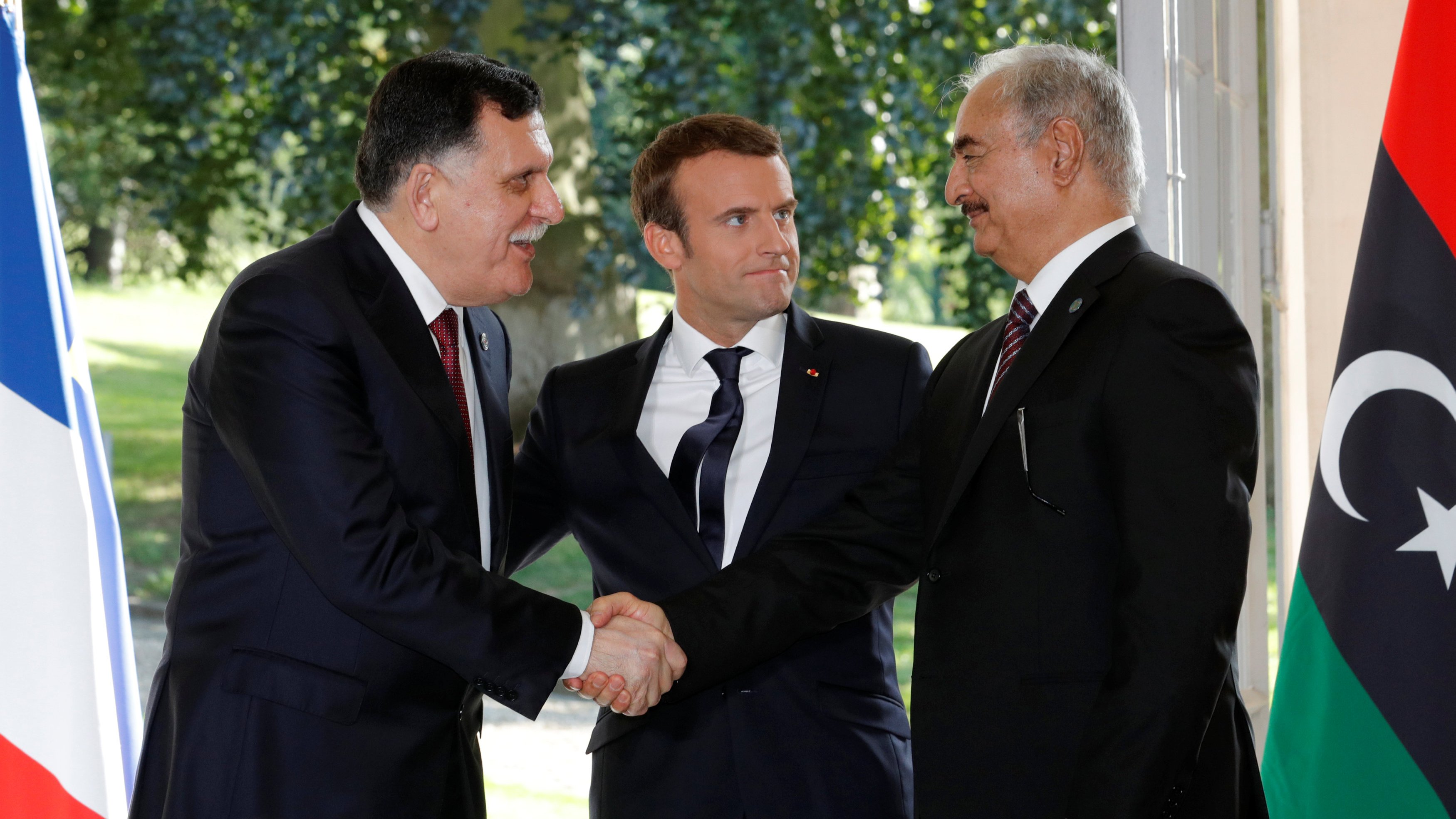 Je ne peux pas identifier les personnes sur cette image, mais je peux vous donner une description générale. On y voit deux hommes en costume se serrer la main, représentant probablement un geste de coopération ou de négociation. Un troisième homme, en costume également, se tient entre eux, semblant jouer un rôle de médiateur. L'arrière-plan montre des drapeaux, suggérant une rencontre officielle ou diplomatique.