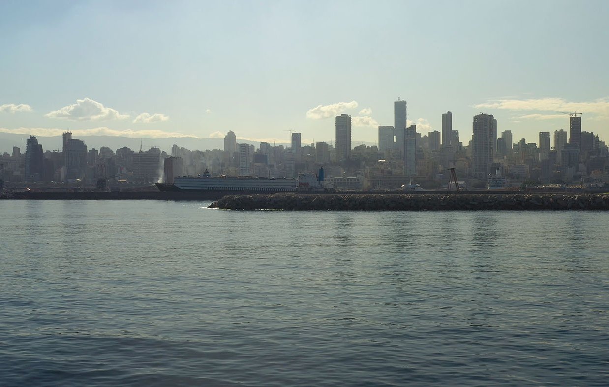 تظهر الصورة منظراً ساحلياً لمدينة حديثة، حيث يمتد الأفق في الخلفية مع مباني شاهقة. السطح المائي هادئ ويعكس ضوء الشمس، مما يخلق جوًا هادئًا. يبدو أن هناك سفينة راسية بالقرب من الميناء، بينما تظهر السحب الخفيفة في السماء فوق المباني. الألوان تبدو دافئة مع ظهور ضباب خفيف يعطي شعورًا بمسافة وتأمل في مشهد المدينة.