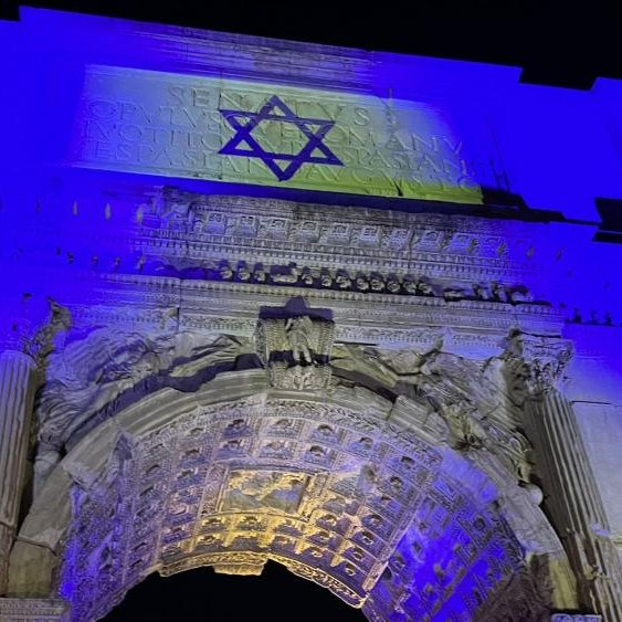 L'immagine mostra un arco a un'altezza notevole, illuminato da luci blu. Sulla parte superiore dell'arco è presente una stella di David, simbolo del giudaismo. L'architettura è riccamente decorata con dettagli ornamentali, evidenziando colonne e incisioni. L'ambientazione notturna conferisce all'opera una presenza suggestiva e solenne.