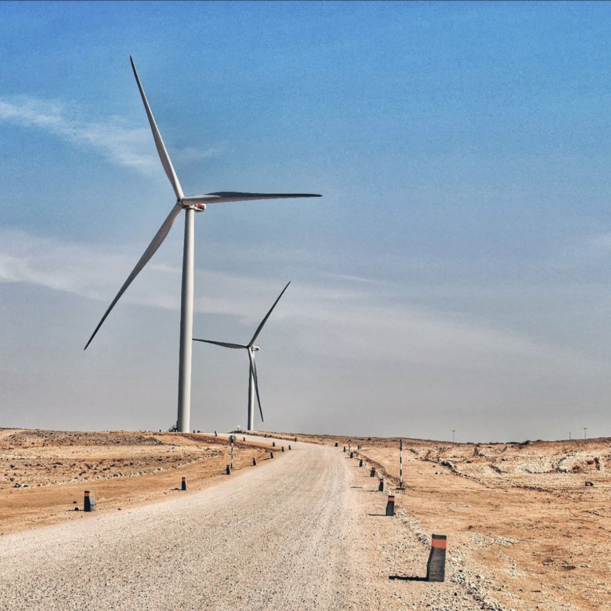 La imagen muestra un paisaje árido y desértico con un camino de grava que se extiende hacia el horizonte. A ambos lados del camino, se pueden ver aerogeneradores, que son grandes turbinas eólicas que aprovechan la energía del viento. El cielo está despejado y presenta un tono azul, lo que sugiere un día soleado. La escena transmite una sensación de tranquilidad y sostenibilidad, con la tecnología eólica destacando en un entorno natural.