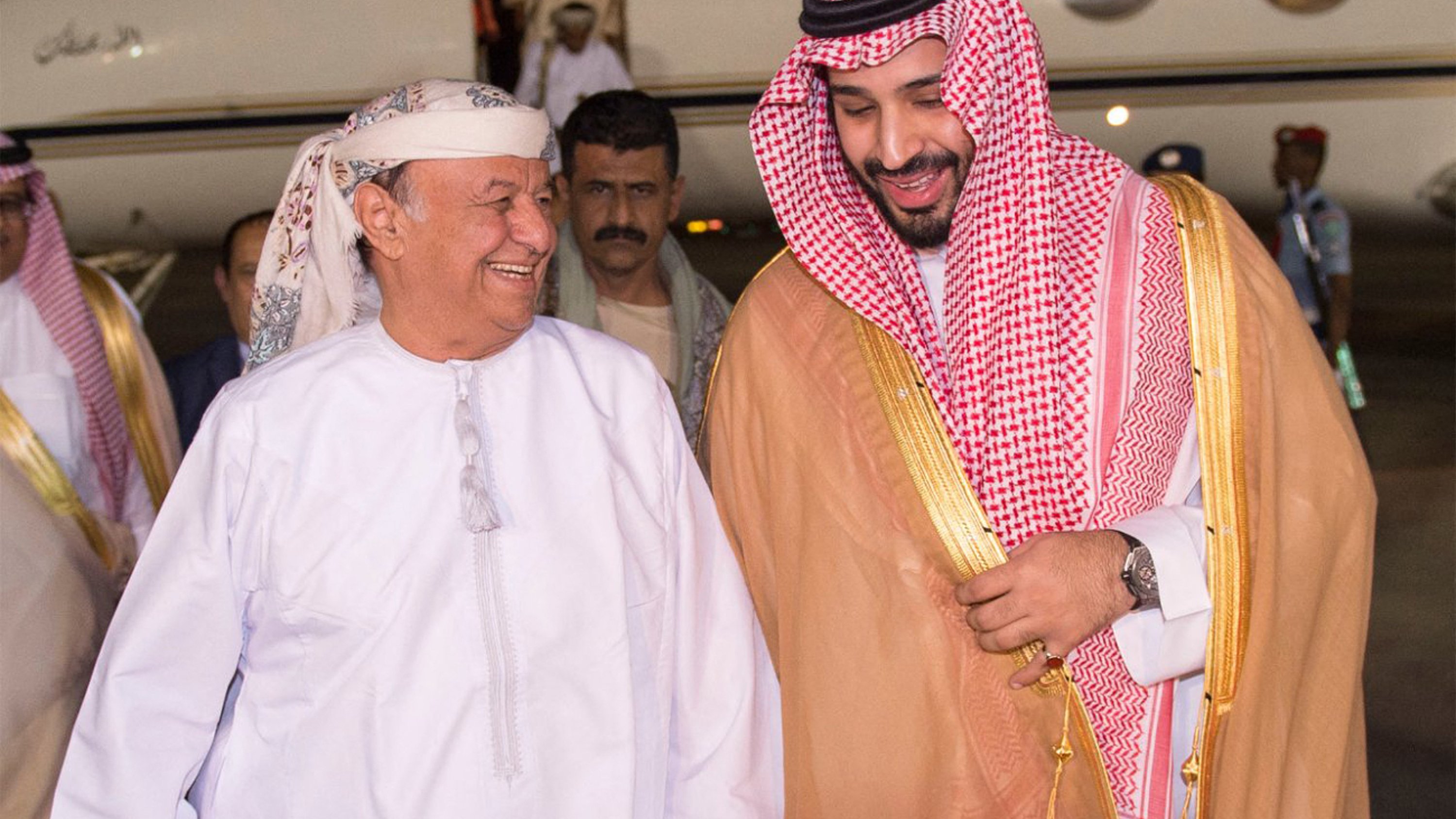 The image shows two men smiling and engaged in a friendly conversation as they walk towards a private jet. One man is dressed in traditional Arab attire, including a white thobe and a headscarf, while the other is wearing a similar traditional outfit, accented with a golden cloak and a checkered headscarf. They appear to be happy and are holding hands, suggesting a warm greeting. In the background, there are several individuals, likely staff or security, standing by the jet. The setting seems to be an airport or a private aviation terminal.