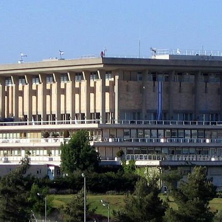 L'image montre un bâtiment moderne avec une architecture distincte, probablement situé dans un environnement urbain. Le bâtiment a plusieurs niveaux, avec des fenêtres et des balcons visibles. On peut également apercevoir des drapeaux, y compris celui d'Israël, flottant en haut du bâtiment. Autour, il y a des arbres et de la verdure, ce qui donne une impression de nature intégrée dans un cadre urbain. Le ciel est bleu et dégagé, ajoutant à la luminosité de la scène.