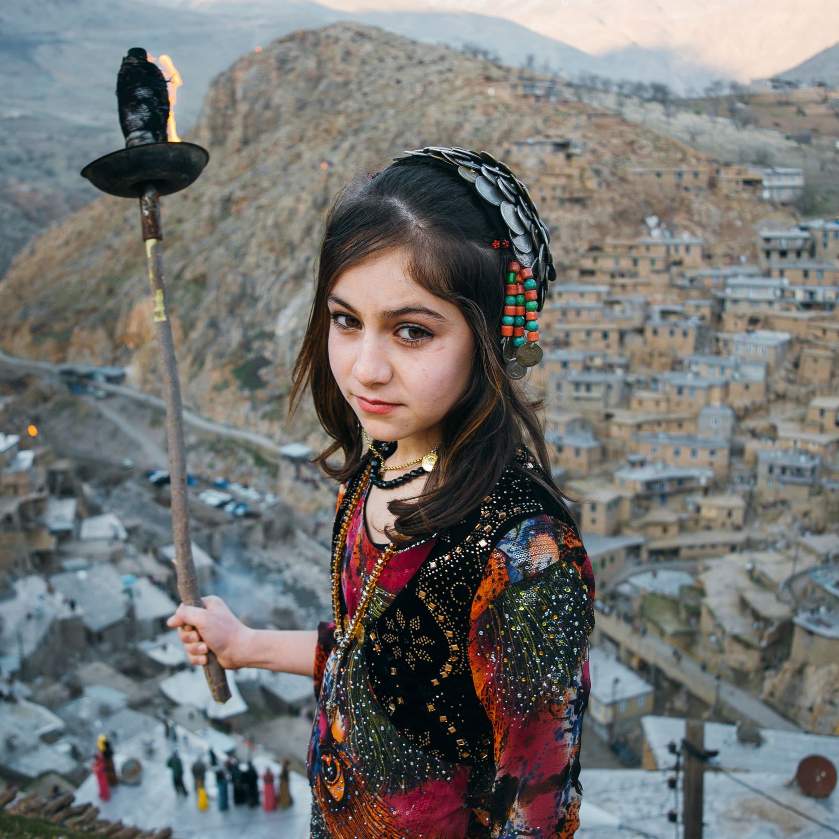 تصویر یک دختر جوان را نشان می‌دهد که در یک محیط کوهستانی و تاریخی ایستاده است. او لباس سنتی رنگارنگی به تن دارد و در دستش مشعل یا شعله‌ای در حال سوختن را نگه‌داشته است. در پس‌زمینه، یک روستای تاریخی با خانه‌های سنگی و دیوارهای کاه‌گلی در دامنه کوه نمایان است. فضای تصویر احساس اصالت و سنت‌های فرهنگی را منتقل می‌کند. دختر با نگاه محکم و جدی به دوربین نگاه می‌کند، که توجه بیننده را جلب می‌کند.