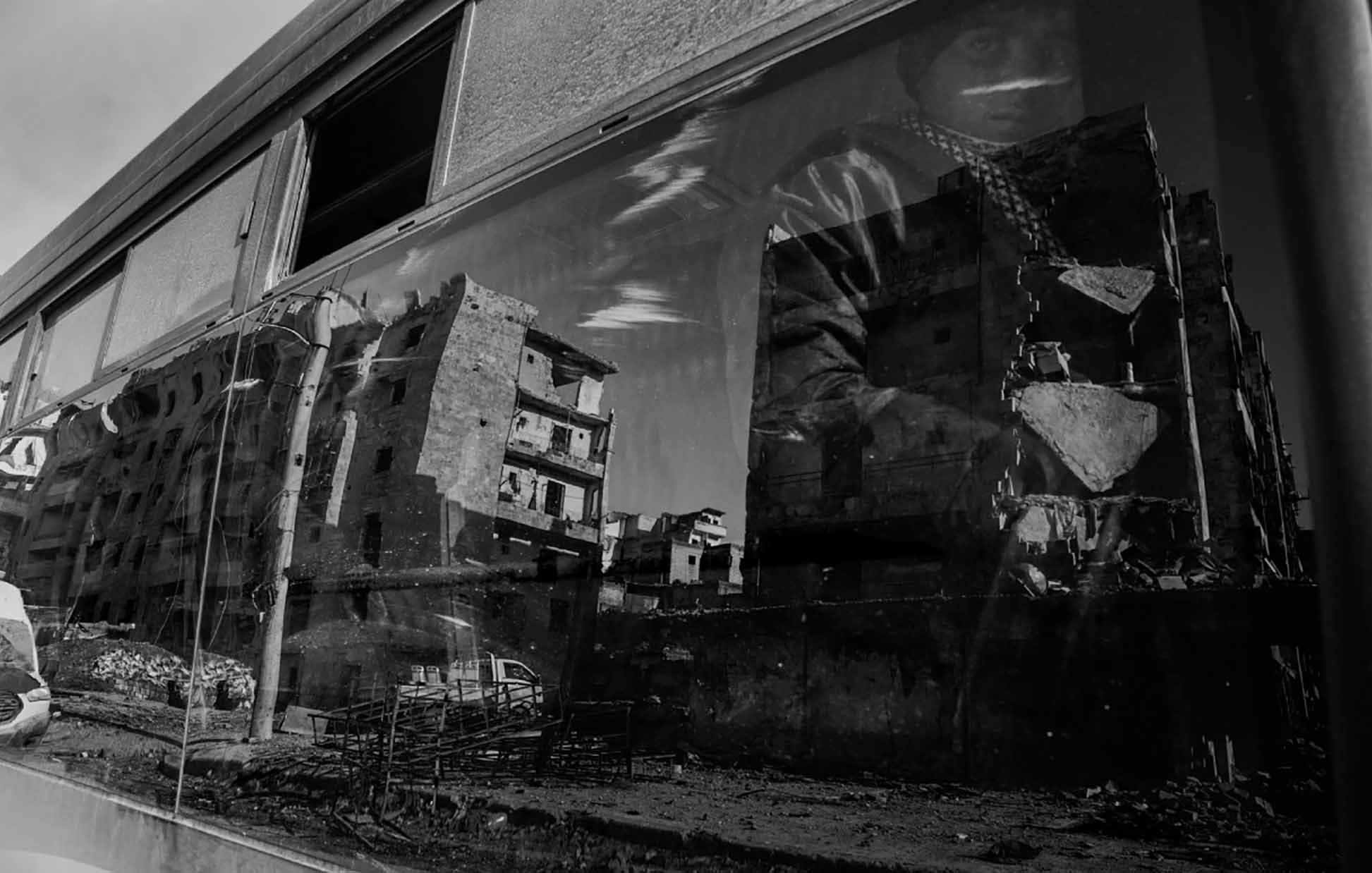 The image features a reflection captured on a glass surface, likely a window or a bus. It shows the distorted but vivid outlines of modern buildings that appear damaged or abandoned, suggesting a scene of urban decay. The reflection also includes a figure partially visible, wearing what looks like a patterned scarf or headdress. The overall tone of the image is monochromatic, emphasizing the somber, evocative nature of the scene. The contrast highlights the interplay between the busy life outside and the desolation depicted in the reflection.
