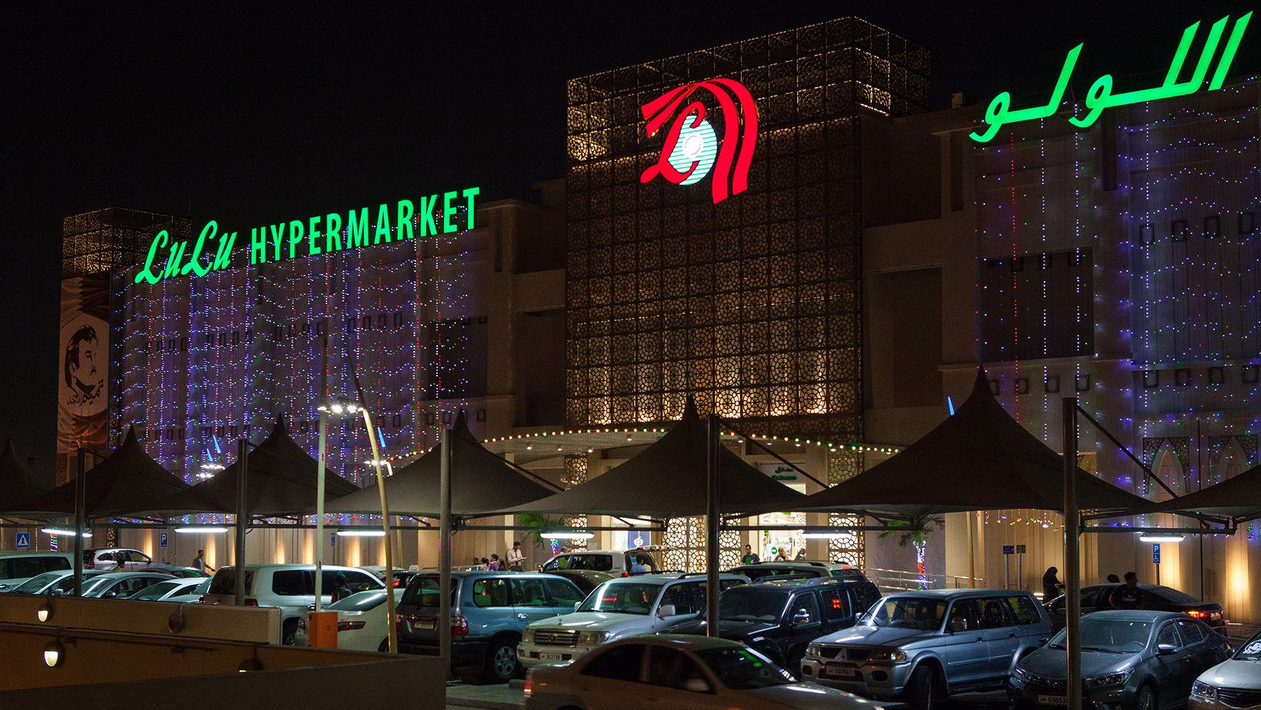 L'image montre un hypermarché, probablement nommé "Lulu Hypermarket", illuminé la nuit. L'extérieur est décoré de lumières, et le nom de l'établissement est affiché en vert, tandis qu'un logo rouge est visible. On aperçoit également un parking rempli de voitures sous des auvents. L'atmosphère paraît animée avec des gens qui semblent entrer ou sortir du magasin.