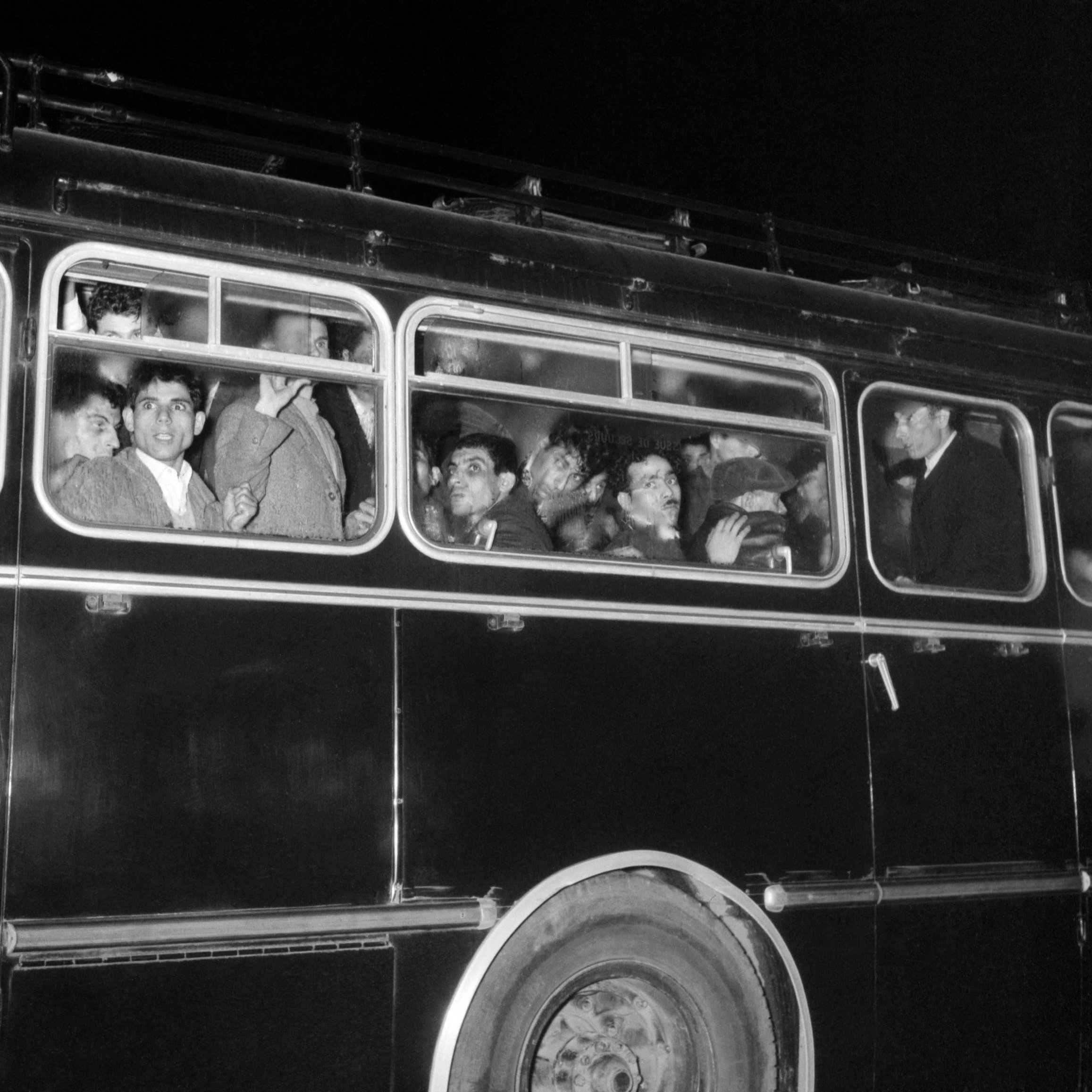 The image depicts a crowded bus at night. Several people are visible through the windows, some looking out while others appear to be engaged in conversation. The bus is dark, possibly painted black, and has a classic design with multiple windows. The expressions of the individuals range from curious to contemplative, highlighting a moment of shared experience among the passengers. The scene captures a sense of movement and typical urban life.