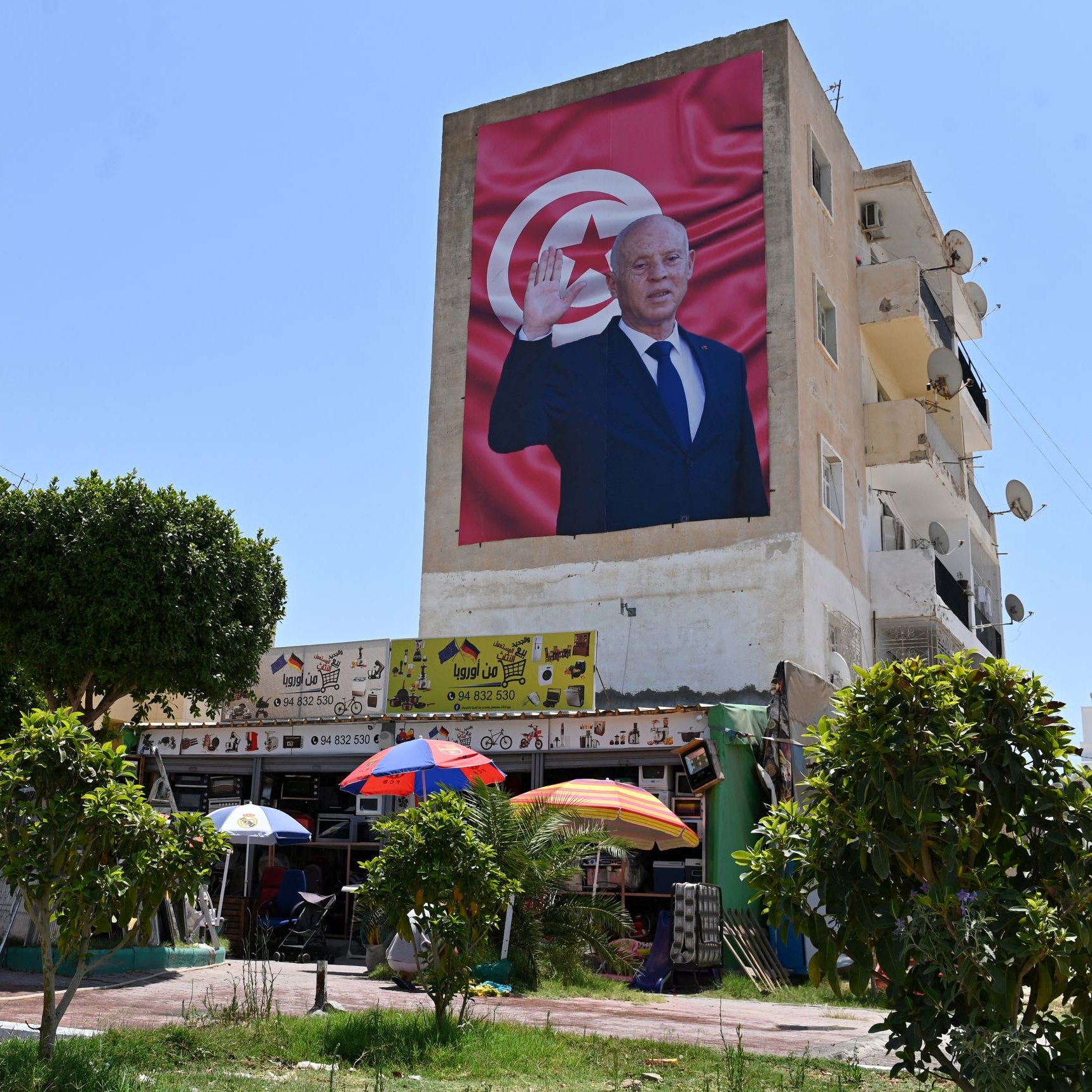 La imagen muestra un gran mural en la pared de un edificio, donde se puede ver a un hombre de pie y saludando. Detrás de él hay una bandera que parece ser de Túnez. En la parte inferior de la imagen, se observa un pequeño mercado o tienda al aire libre, con sombrillas de colores y algunas personas. Hay árboles y bicicletas en el entorno, lo que sugiere un ambiente urbano y concurrido. El cielo está despejado y soleado.