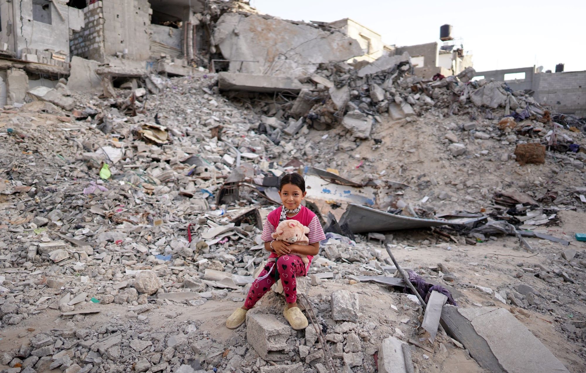L'image montre une jeune fille assise sur des décombres, entourée de ruines. Elle porte des vêtements colorés et tient un jouet, semblant à la fois vulnérable et résiliente au milieu du chaos. La scène évoque des sentiments de tristesse et de perte, en raison de la destruction visible sur son environnement.