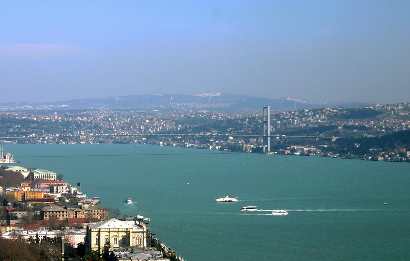 تصویر نمای کلی از یک شهر در کنار یک دریاچه یا رودخانه را نمایش می‌دهد. در سمت چپ تصویر، ساختارها و ساختمان‌های مختلفی دیده می‌شود که نشان‌دهنده مناطق مسکونی و تجاری است. در پس‌زمینه، پل بزرگی وجود دارد که دو طرف آب را به هم متصل می‌کند. آسمان آبی و صاف است و دورنمای تپه‌ها و شهر در پس‌زمینه به وضوح قابل مشاهده است. همچنین، برخی قایق‌ها در سطح آب در حال حرکت هستند. این تصویر حس سرزندگی و زیبایی طبیعی منطقه را منتقل می‌کند.