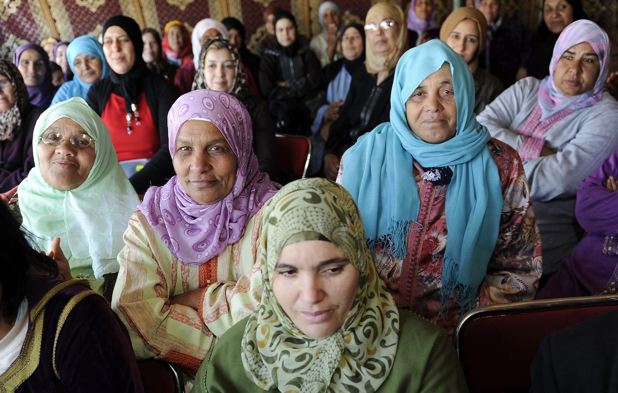 La imagen muestra a un grupo de mujeres, la mayoría de ellas vestidas con hijabs de diferentes colores y estilos. Ellas están sentadas en un ambiente que parece ser un evento o reunión, y sus expresiones varían entre la seriedad y la atención. Algunas mujeres llevan prendas tradicionales, y el fondo sugiere un entorno decorado con motivos culturales. La diversidad en los colores de los pañuelos y vestimenta resalta un sentido de unidad y diversidad entre ellas.