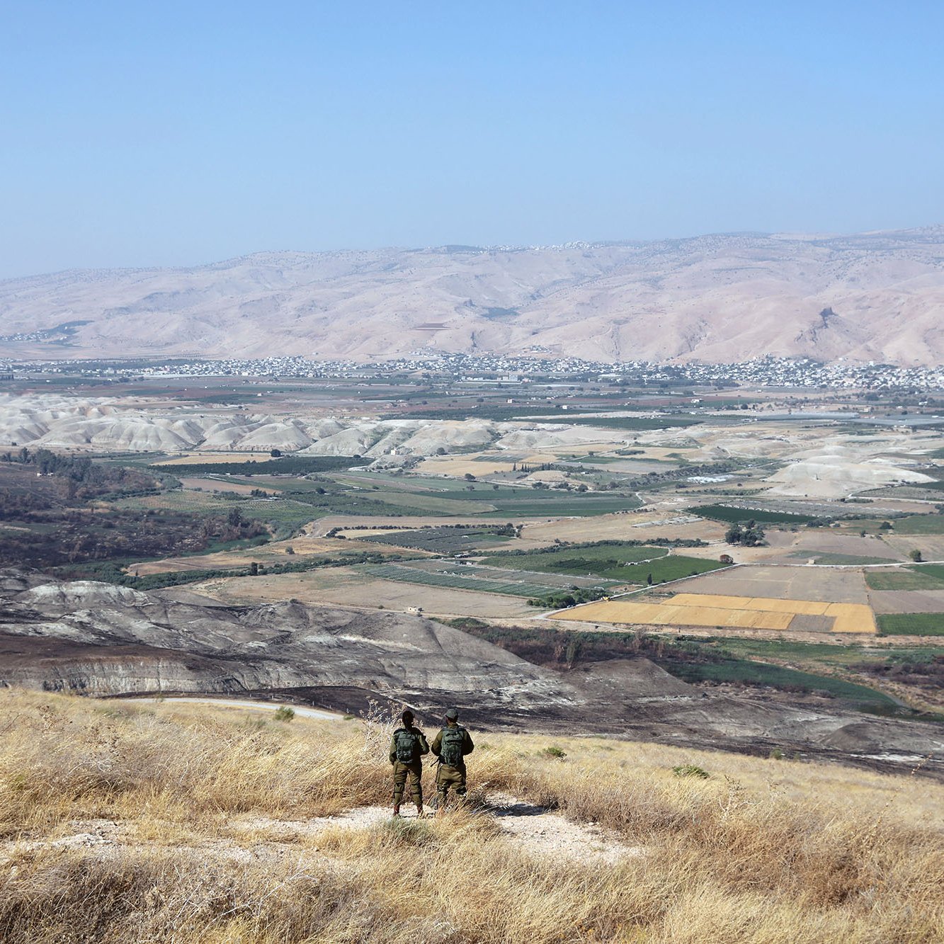La imagen muestra un paisaje vasto y montañoso, con colinas y valles amplios. En primer plano, se pueden ver dos personas de pie, posiblemente soldados, observando el paisaje desde una cima. El área está cubierta de hierba seca y destaca un amplio valle con campos de cultivo en diferentes tonos de verde y amarillo. En el fondo, se aprecian montañas que se extienden hasta el horizonte bajo un cielo despejado.