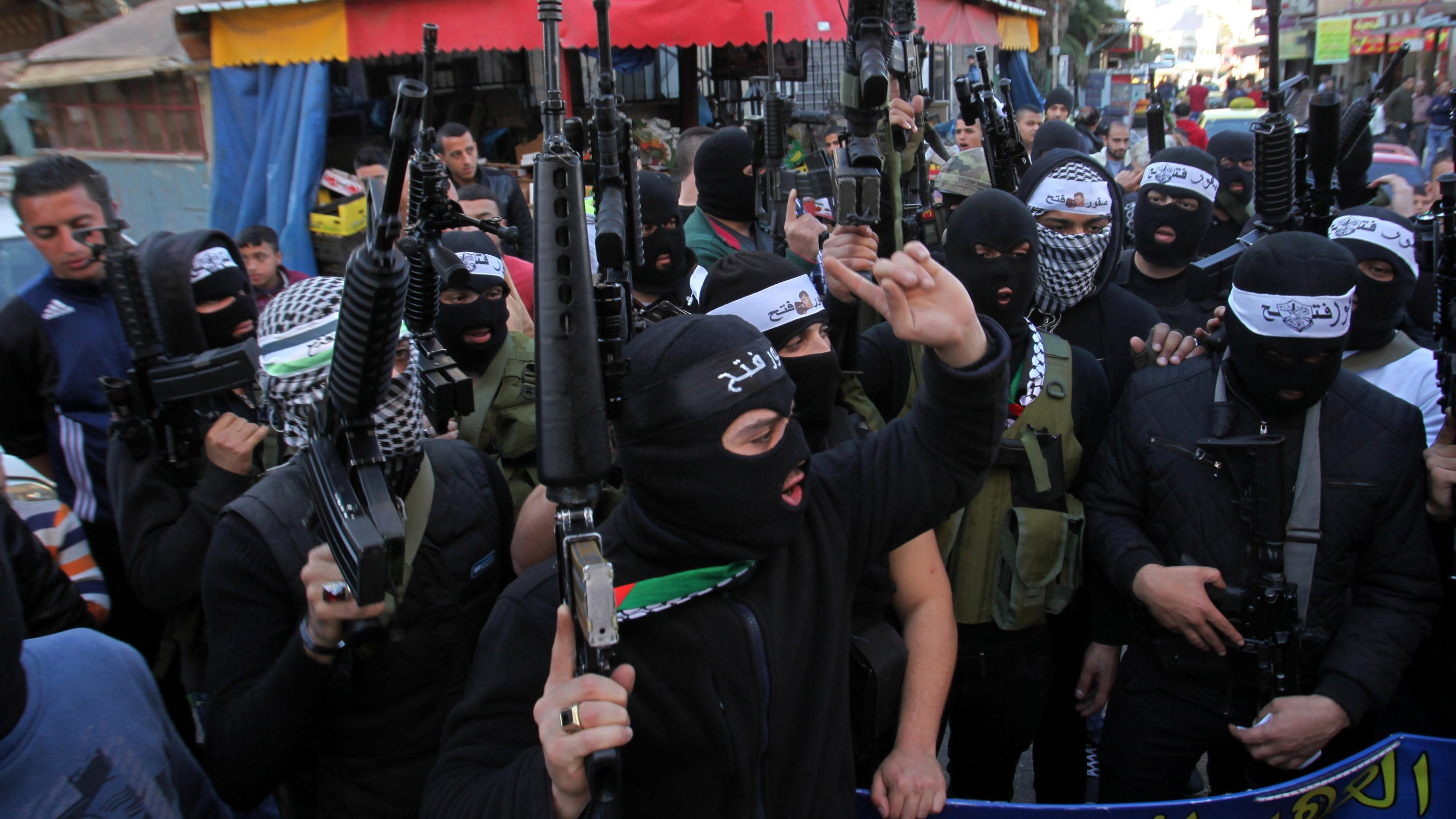 L'image montre un groupe de personnes portant des masques noirs et tenant des armes. Elles se rassemblent dans une rue, avec des bâtiments et des commerçants en arrière-plan. L'atmosphère semble tendue, et leurs expressions indiquent une certaine détermination ou colère. Des drapeaux et des banderoles sont visibles, suggérant un contexte de manifestation ou de rassemblement politique.