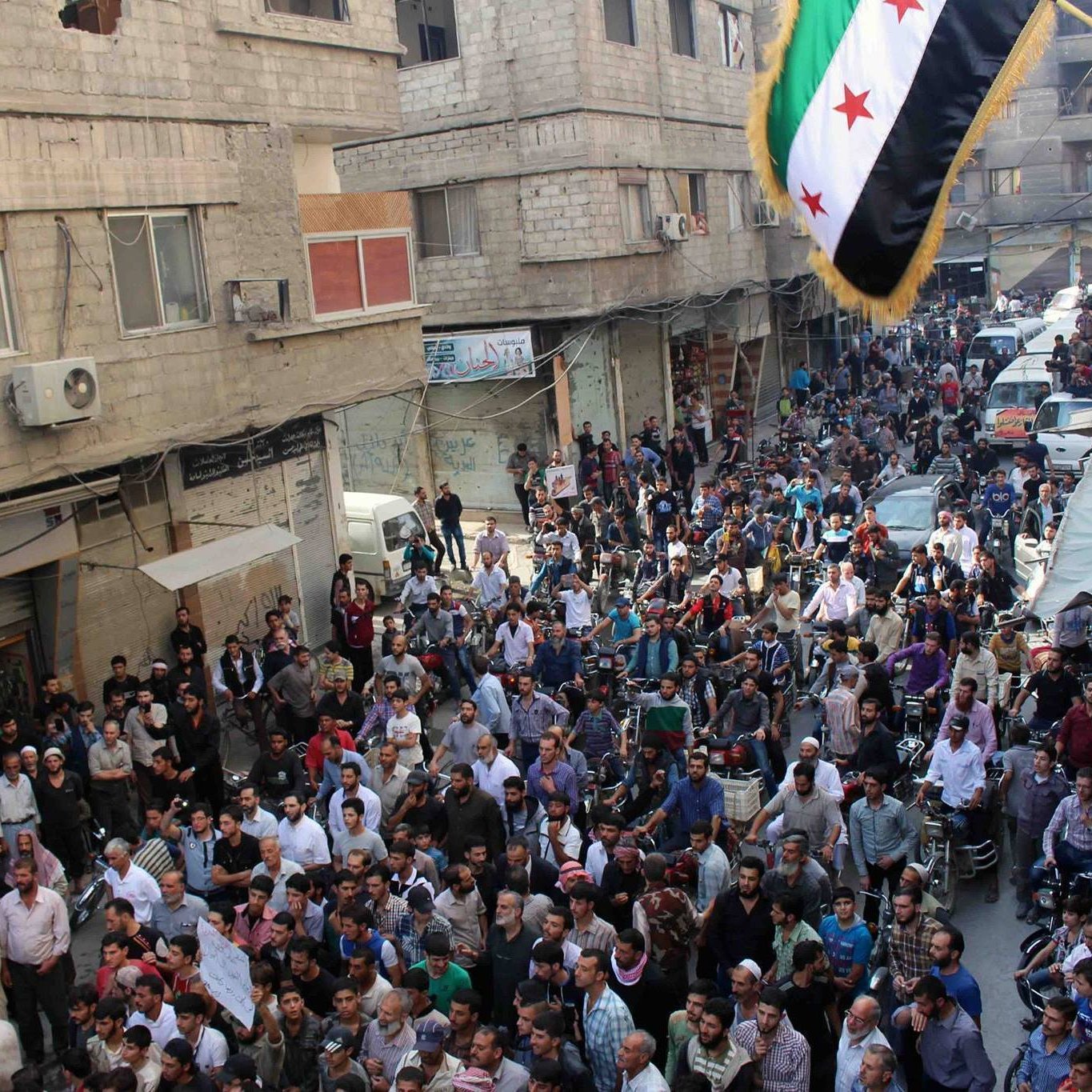 L'image montre une grande foule rassemblée dans une rue, probablement dans une ville syrienne. Les gens semblent se réunir pour une manifestation ou un événement public, avec des pancartes et un drapeau aux couleurs vives, typiquement associé à des mouvements de protestation. Les bâtiments environnants affichent des signes de vie urbaine, et plusieurs véhicules sont stationnés dans la rue. L'atmosphère semble intensément animée, avec une forte concentration de personnes dans l'espace.