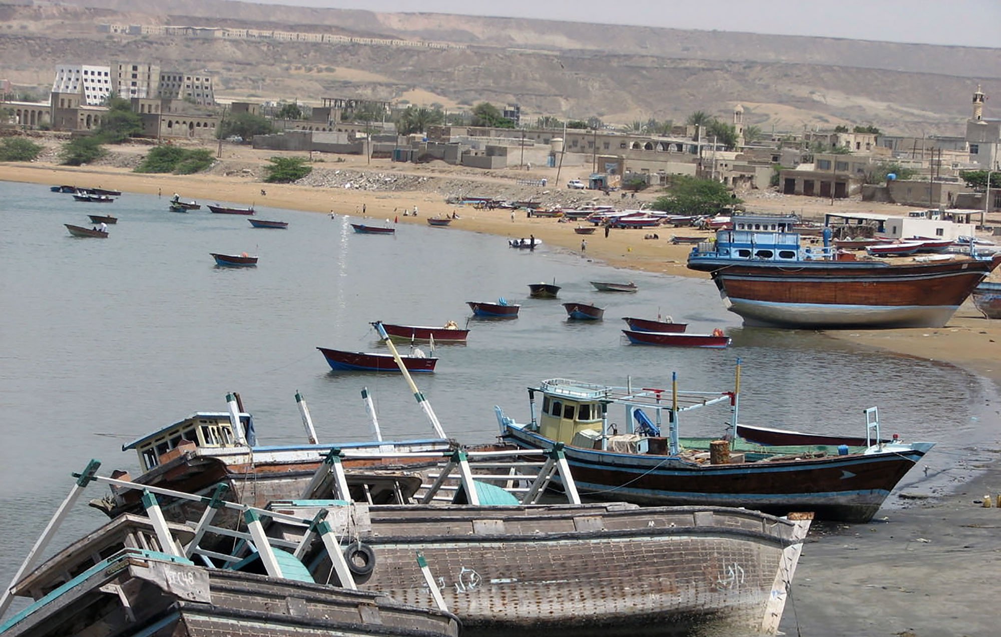 The image depicts a coastal scene with several fishing boats moored or resting on the shore. Some boats are partially submerged in shallow water, while others are docked along the beach. The background features sandy shores and low-lying buildings, with a mountainous area visible in the distance. The overall atmosphere suggests a tranquil seaside environment, typical of a fishing village.