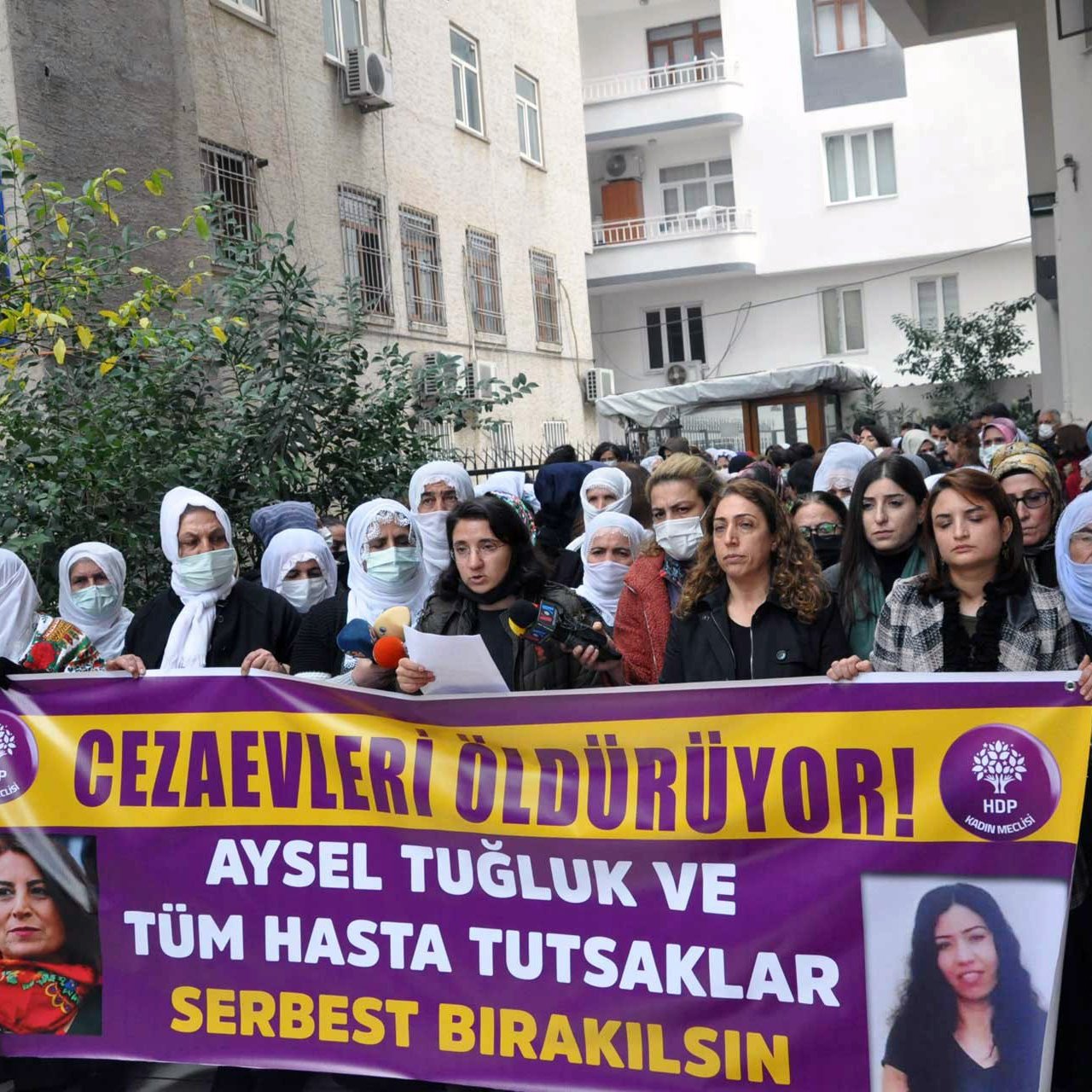 En la imagen, un grupo de personas se encuentra reunido en una manifestación. Hay mujeres con pañuelos en la cabeza, y algunas de ellas sostienen pancartas que expresan mensajes en apoyo a Aysel Tuğluk y a otros prisioneros enfermos. El ambiente parece ser de protesta, con un enfoque en la liberación de detenidos. Se observa una edificación al fondo, que sugiere que la manifestación se lleva a cabo frente a un edificio público o institucional.