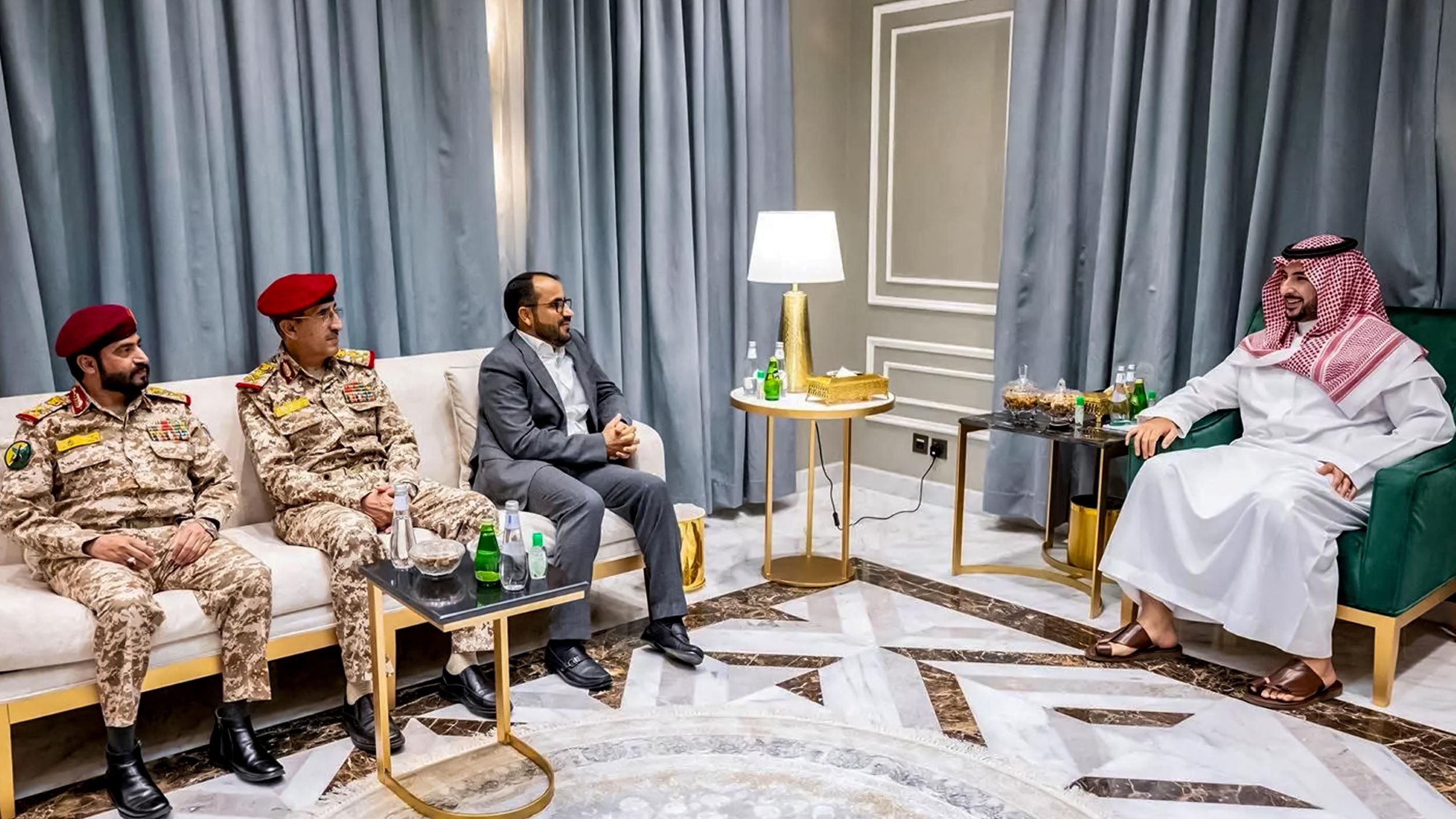 تصویر یک جلسه رسمی را نشان میدهد. در این تصویر چهار مرد در یک محیط داخلی نشستهاند. دو نفر از آنها لباس نظامی با کلاههای قرمز به تن دارند و به نظر میرسند که در مقامهای نظامی هستند. نفر سومی که لباس رسمی دارد، در وسط نشسته و به نظر میرسد در حال گفتگو با شخص چهارمی است که لباسی سنتی عربی به تن دارد. این شخص چهارم در طرف راست تصویر نشسته و به دیگران توجه دارد. محیط دارای دکوراسیون مدرن و مبلمان راحتی است. همچنین یک لامپ روشن و میزهایی با نوشیدنیهایی روی آنها مشاهده میشود.