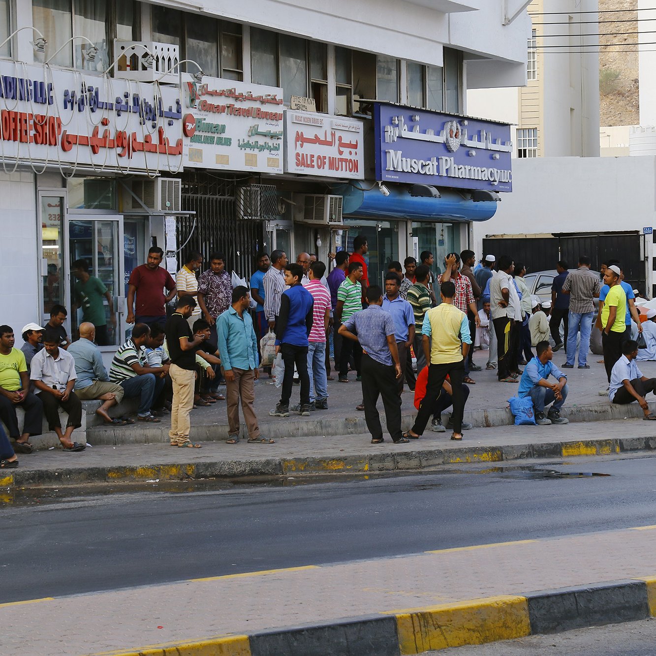 تظهر الصورة مشهداً من شارع في منطقة حضارية مزدحمة. يوجد العديد من الأشخاص يتجمعون بالقرب من storefrontات ومتاجر متنوعة. يمكن رؤية مجموعة من الرجال والمارة يجلسون أو يقفون، وبعض الناس مشغولون بالتحدث مع بعضهم. يضم المشهد أيضاً لافتات باللغة العربية تشير إلى المتاجر والمرافق. الجو يبدو نشطًا، مع حركة مرور على الشارع في الخلفية.