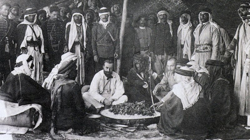 L'image montre un groupe de personnes, probablement issus de cultures arabes, rassemblées autour d'un grand plat de nourriture posé au sol. Certains sont assis tandis que d'autres se tiennent debout en arrière-plan. Les participants portent des vêtements traditionnels, tels que des robes et des turbans, ce qui suggère un contexte historique. L'ambiance semble conviviale, et l'événement pourrait être un repas ou une célébration. Des éléments du décor indiquent que la scène se déroule en extérieur, possiblement sous une tente.