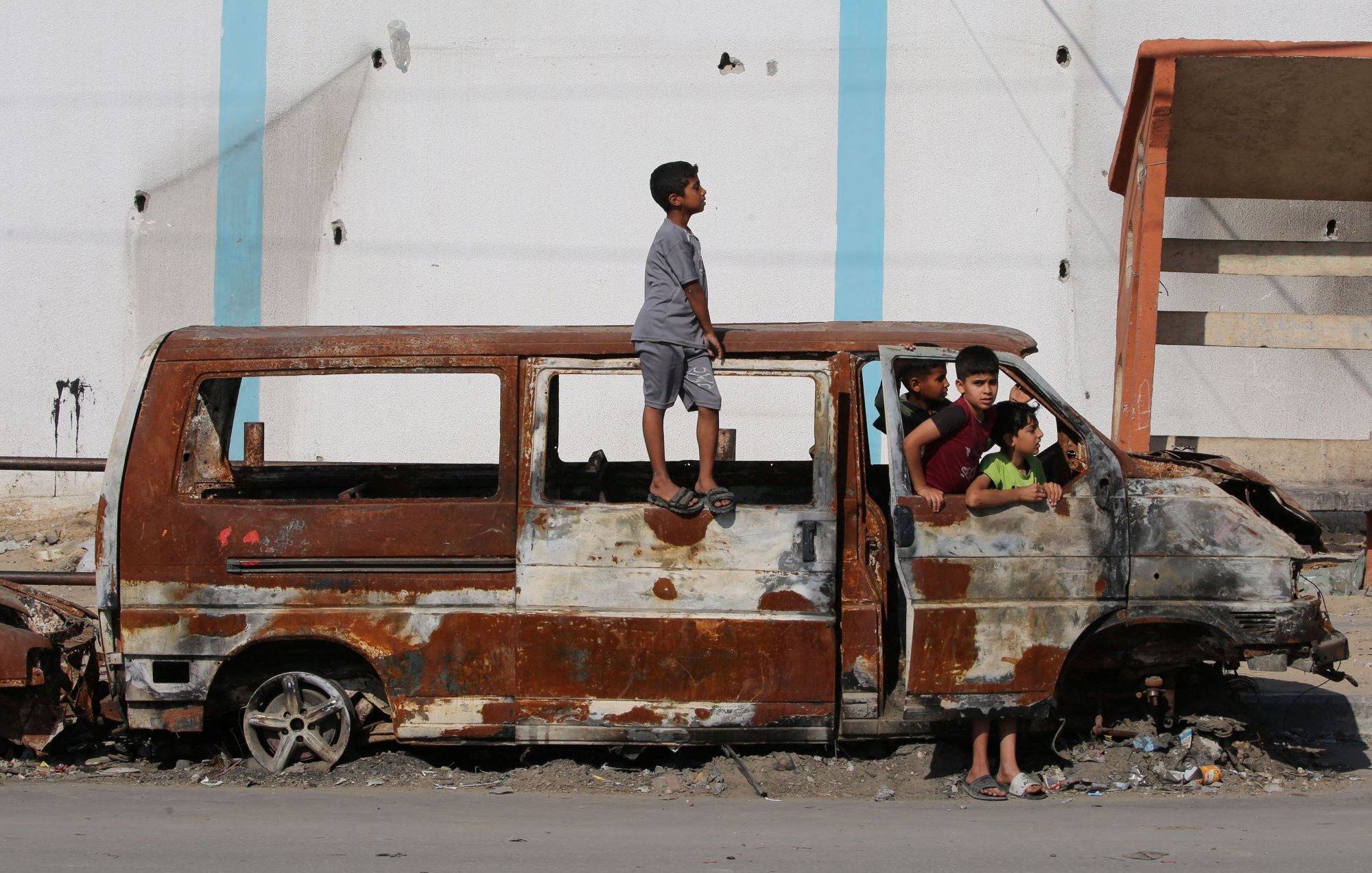 L'image montre un groupe d'enfants jouant autour d'une vieille fourgonnette rouillée et endommagée. Un enfant est debout sur le toit de la voiture, tandis que d'autres sont assis et debout autour d'elle, semblant s'amuser malgré l'environnement délabré. En arrière-plan, on aperçoit un bâtiment aux murs abîmés. L'atmosphère semble à la fois joyeuse et nostalgique, illustrant la résilience des enfants dans un cadre difficile.