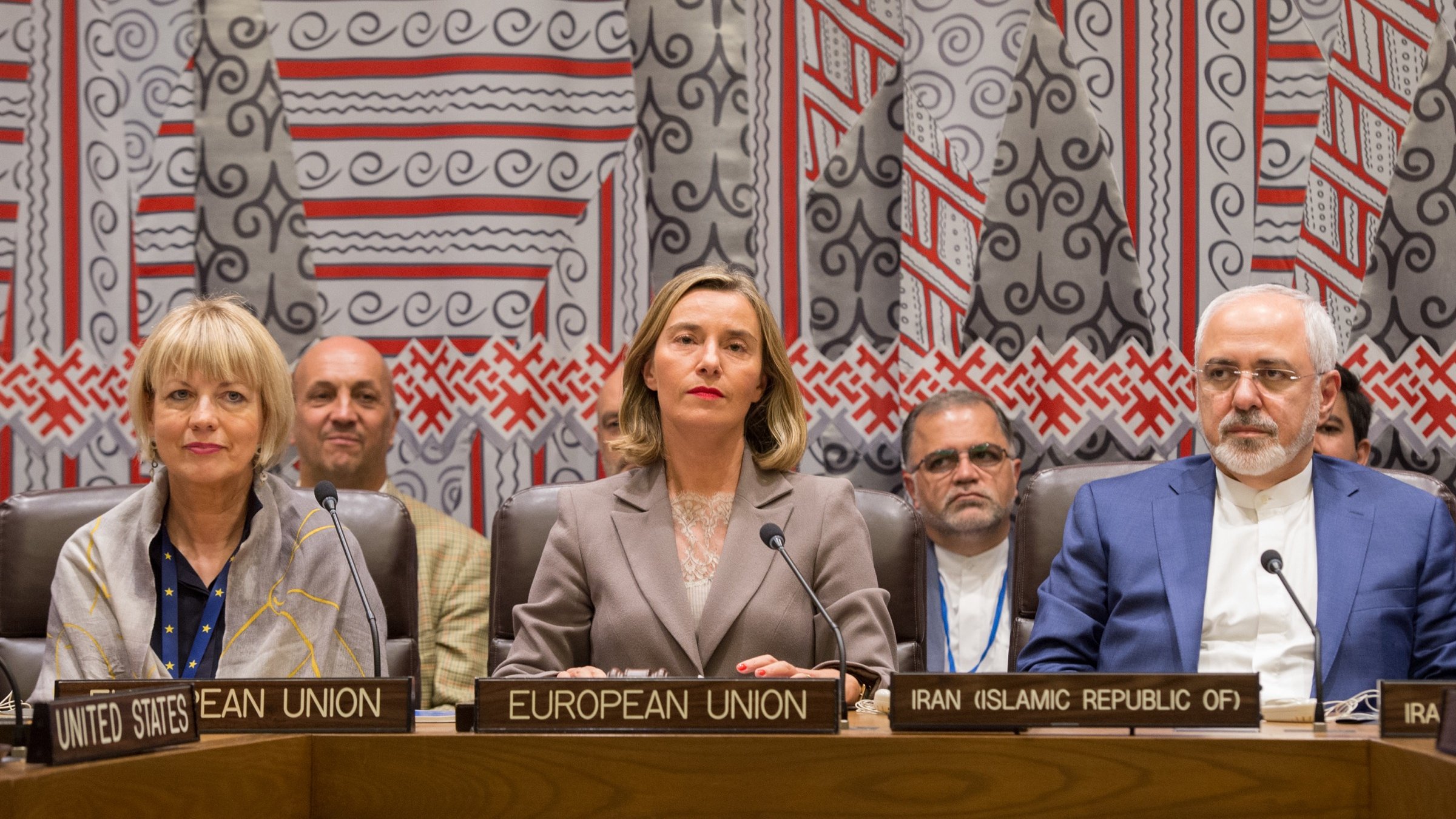 L'image montre une réunion internationale avec plusieurs personnes assises à une table. On peut voir des panneaux identifiant les différents participants, tels que l'Union européenne et l'Iran. Trois figures centrales semblent être des représentants importants, avec des expressions sérieuses. En arrière-plan, il y a des individus qui écoutent attentivement. Le décor est riche et coloré, avec des motifs géométriques sur le mur derrière eux, suggérant un cadre formel pour des discussions diplomatiques.