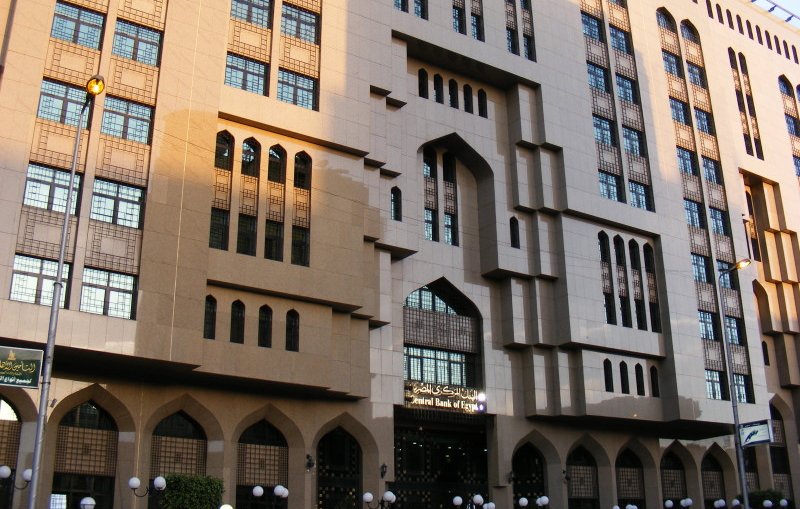 L'image montre un bâtiment architectural impressionnant, avec une façade en pierre et de grandes fenêtres ornées. Les éléments décoratifs évoquent un style inspiré de l'architecture islamique, notamment avec des arches et des motifs géométriques distincts. La lumière du soleil met en valeur les détails de la structure, tandis que des lampadaires et des lampes décoratives sont visibles au premier plan. Cela dégage une ambiance à la fois moderne et traditionnelle.