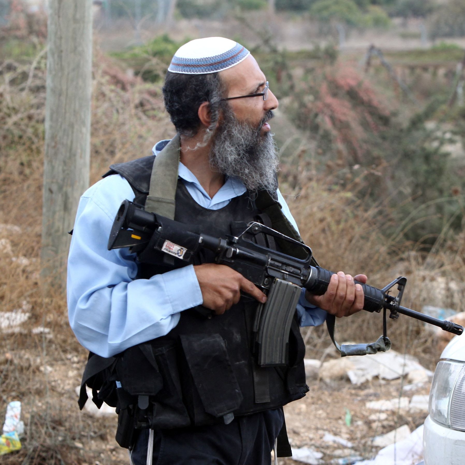 L'immagine mostra un uomo in piedi in un ambiente all'aperto con vegetazione secca sullo sfondo. Indossa un kippah e un giubbotto tattico, e tiene in mano un fucile. L'atteggiamento e l'abbigliamento suggeriscono un contesto di sicurezza o di conflitto. Accanto a lui si vede un veicolo. La scena sembra essere in una zona rurale o periferica.