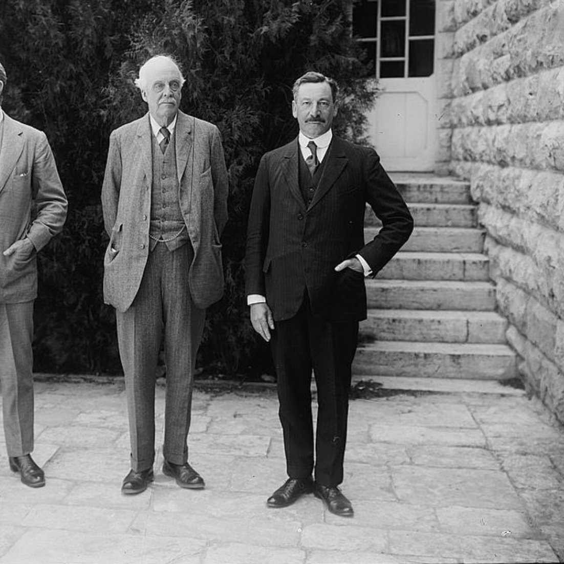 سه مرد در لباس رسمی ایستاده‌اند. پس‌زمینه شامل درختان و یک سازه سنگی ساده است.