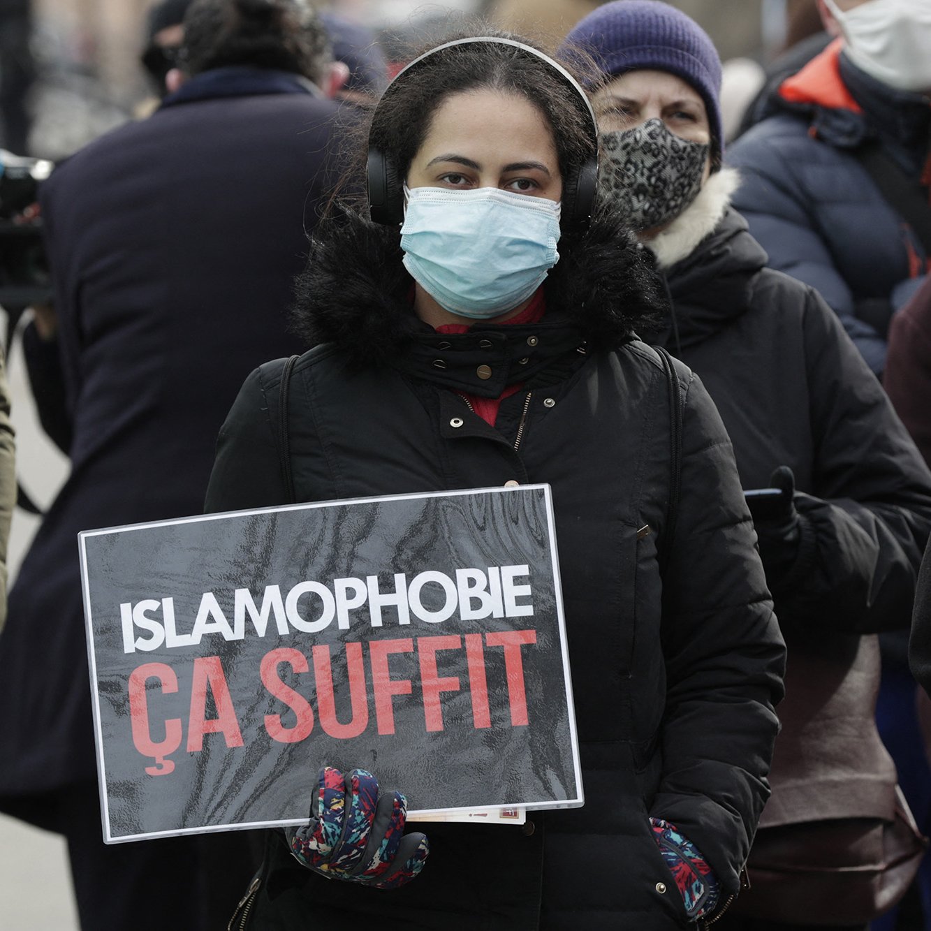 En la imagen se puede ver a una persona sosteniendo un cartel que dice "ISLAMOPHOBIE ÇA SUFFIT", lo que se traduce como "ISLAMOFOBIA BASTA YA". La persona lleva puesta una mascarilla y parece estar en un evento o manifestación, rodeada de otras personas que también usan mascarillas. El ambiente sugiere una expresión de protesta o de apoyo a una causa relacionada con la lucha contra la islamofobia.