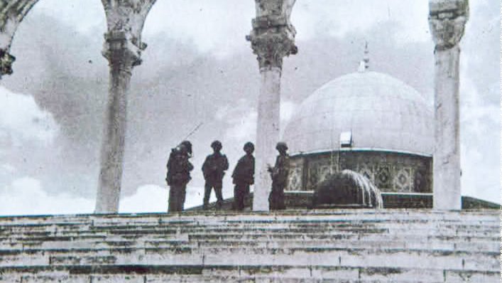 L'image montre une scène en noir et blanc, probablement historique, avec un groupe de soldats se tenant sur des marches devant un édifice. On peut apercevoir un dôme caractéristique au fond, ainsi que des arches en pierre en avant-plan. L'atmosphère est solennelle et le lieu semble chargé d'histoire. Les soldats sont habillés de manière militaire et semblent en position de garde.