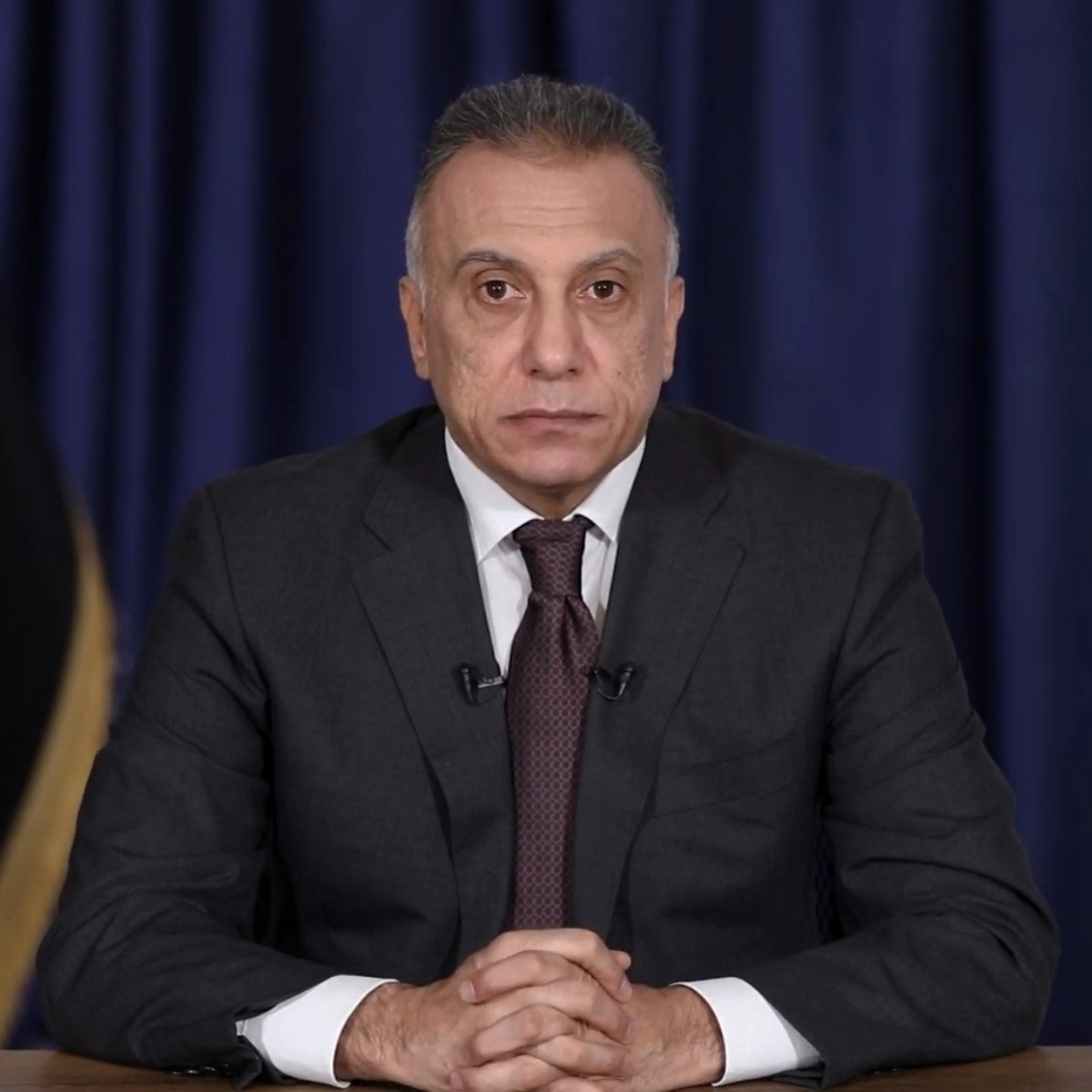 L'image montre un homme assis à une table, vêtu d'un costume sombre et d'une cravate. Il a une expression sérieuse et regarde directement l'objectif. En arrière-plan, il y a un rideau bleu et un drapeau irakien, ce qui suggère qu'il se trouve dans un cadre officiel ou politique.