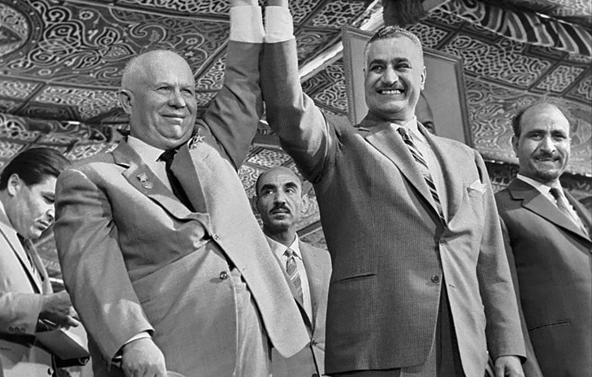 L'image montre deux hommes se tenant la main, souriant et levant les bras en signe de victoire ou de camaraderie. Ils sont entourés de plusieurs autres hommes en arrière-plan, qui semblent également participer à l'événement. Les tenues des protagonistes suggèrent un contexte formel ou officiel, avec des costumes élégants. Le décor en arrière-plan présente des motifs ornementaux, indiquant un événement culturel ou politique. L'ambiance générale semble être celle d'une célébration ou d'une réunion importante.