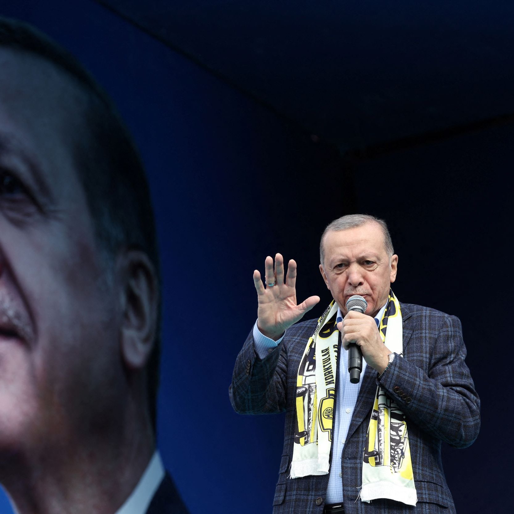 Nell'immagine si vede un uomo che parla a un pubblico, indossando una giacca a quadri e una sciarpa. Dietro di lui c'è un grande murale o un'immagine di un volto che lo ritrae. L'ambientazione sembra essere un evento pubblico o un comizio, con un'atmosfera di fervore e partecipazione. Il fondo è di colore blu, creando un contrasto con il soggetto principale.