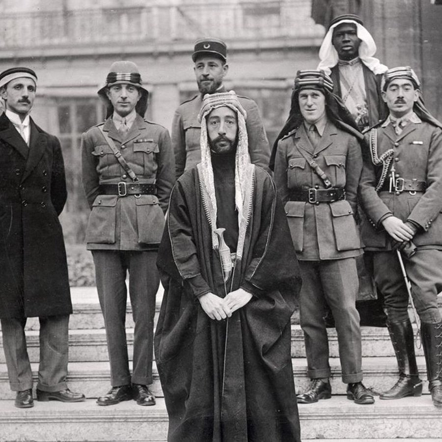 L'image montre un groupe de six hommes posant ensemble. Ils sont vêtus de différentes tenues militaires et traditionnelles. Au centre, un homme en robe traditionnelle, portant une tête couronnée, semble être le principal sujet. À sa gauche, un autre homme en uniforme militaire. Les autres sont également en uniformes variés, indiquant peut-être des origines et des rôles différents. L'arrière-plan montre une architecture qui semble historique. L'ensemble dégage une atmosphère de confluence culturelle et de collaboration.