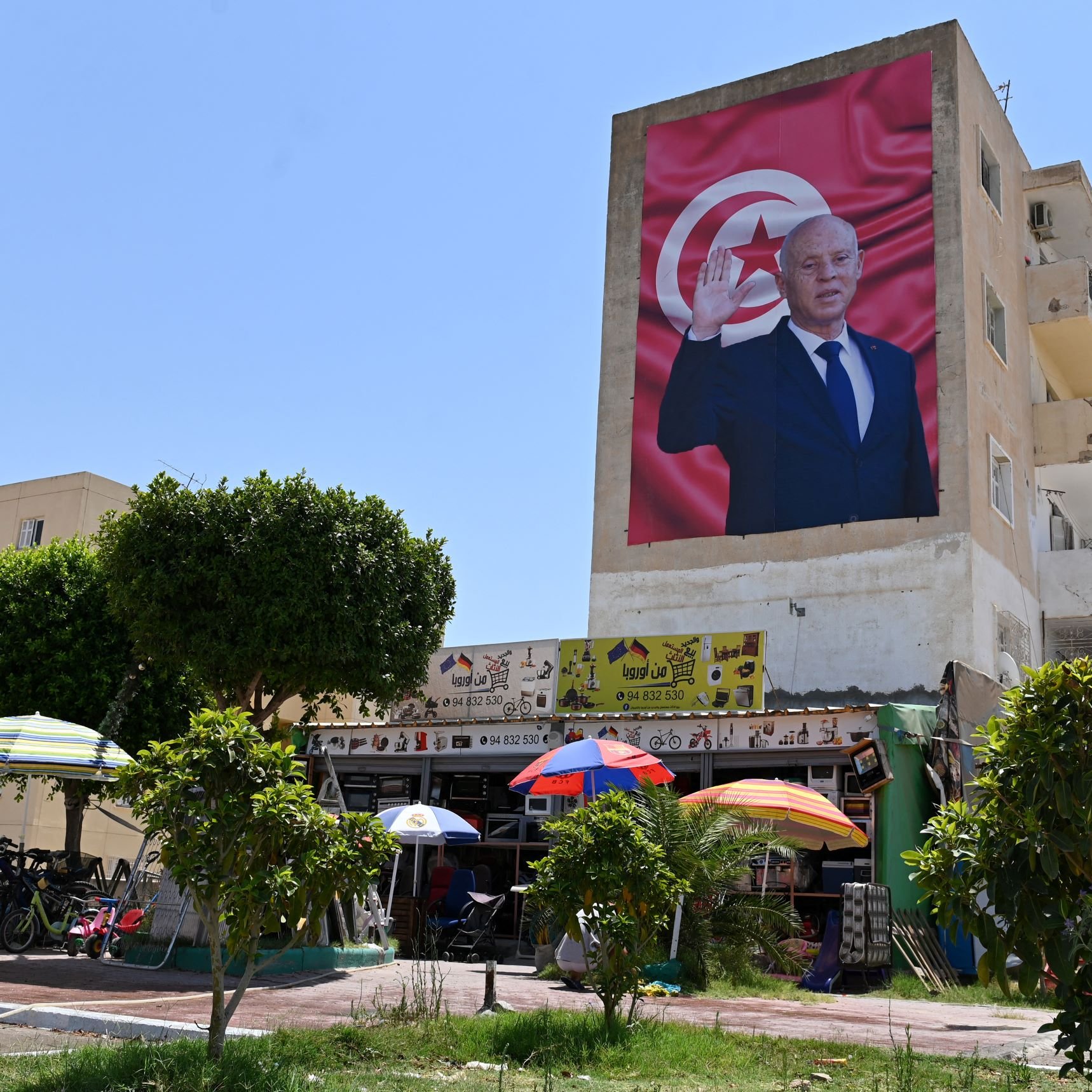 L'immagine mostra un grande murales su un edificio, raffigurante una figura maschile che saluta con una mano alzata. Sullo sfondo, è presente una bandiera che presenta i colori e il simbolo della Tunisia. Nella parte inferiore si possono vedere alcuni ombrelloni colorati, accanto a un'area con biciclette e persone, il tutto immerso in un ambiente urbano. Il cielo è sereno e luminoso, suggerendo una giornata di sole.