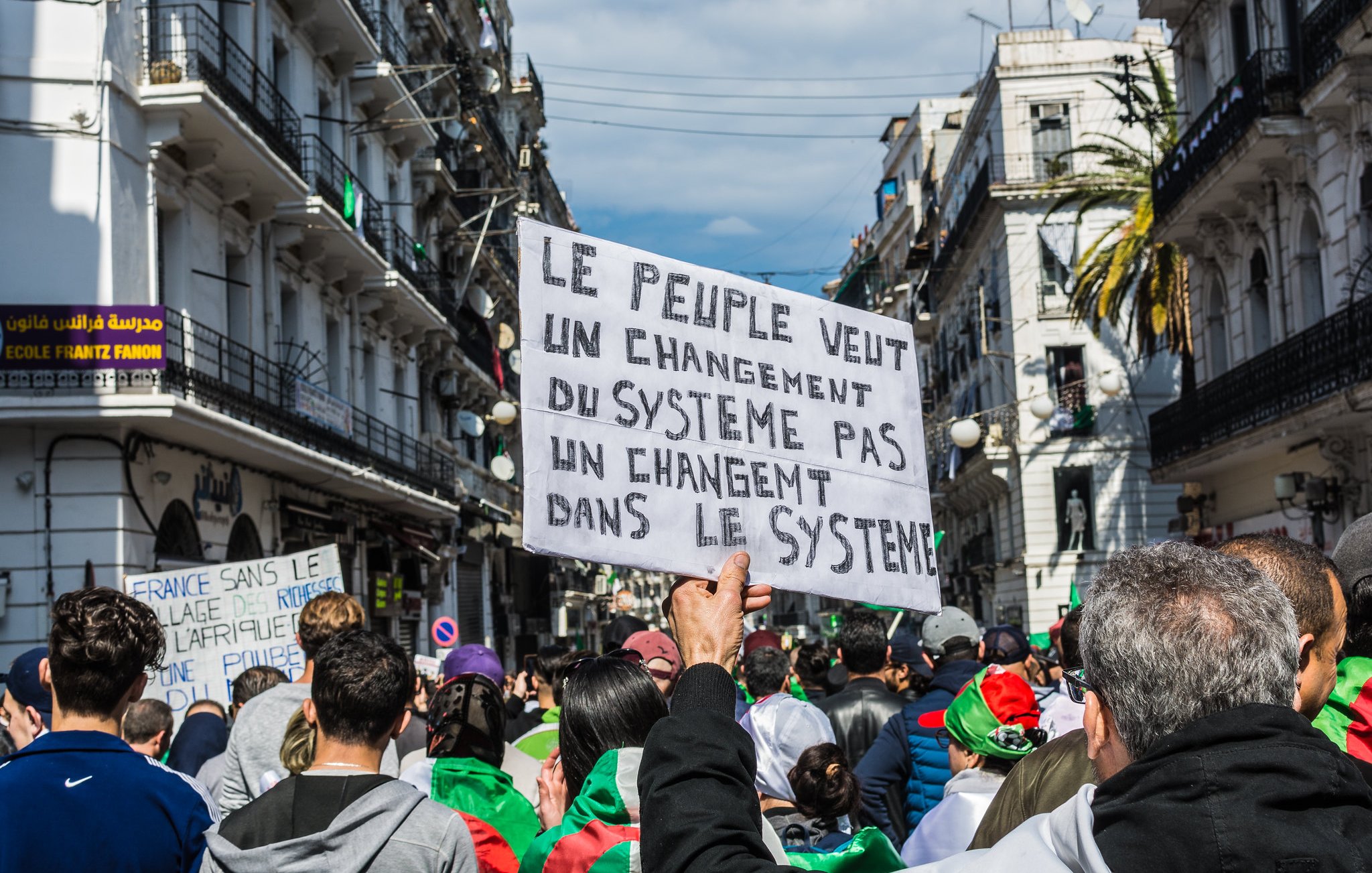 L'image montre une manifestation dans une rue animée. Au premier plan, une personne tient une pancarte qui exprime un message clair : "Le peuple veut un changement du système pas un changement dans le système". On aperçoit des manifestants qui portent des drapeaux et semblent mobilisés. Les bâtiments environnants évoquent une ambiance urbaine et dynamique, tandis que le ciel est partiellement nuageux. L'ensemble traduit une forte volonté collective de changement.