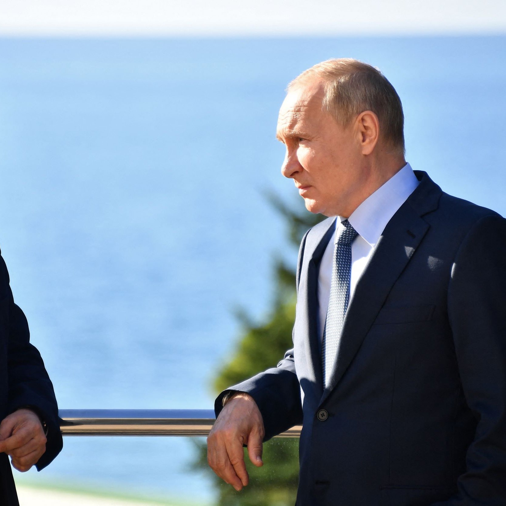 در این تصویر، دو مرد در حال گفتگو هستند. یکی از آن‌ها کت و شلوار تیره به تن دارد و به سمت دیگر شخص اشاره می‌کند. مرد دیگر نیز کت و شلوار رسمی پوشیده و به سمت شخص اول نگاه می‌کند. پس‌زمینه تصویر نیز شامل دریا و آسمان است که فضایی آرامش‌بخش را ایجاد کرده است.