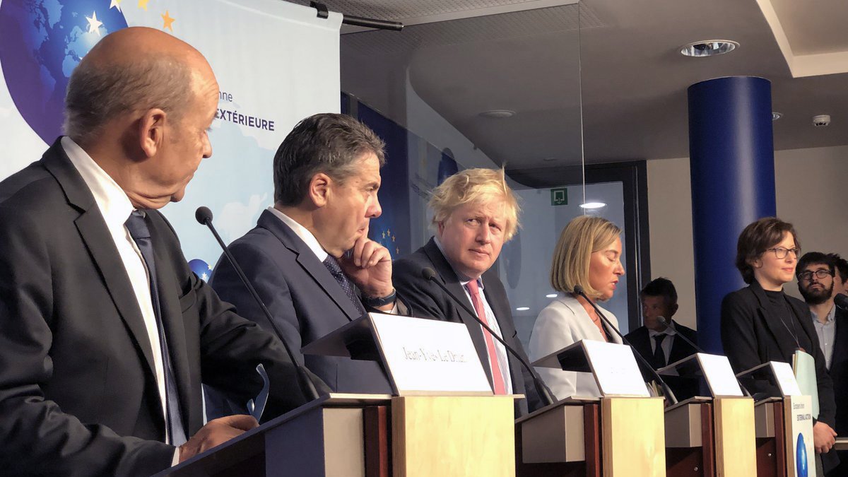 در این تصویر، گروهی از سیاستمداران در حال برگزاری یک کنفرانس خبری یا نشست مطبوعاتی هستند. چندین نفر بر روی چهار پایه ایستاده‌اند که هر کدام به یک میکروفن مجهز هستند. در میان آن‌ها مردی با موهای بلوند و کوتاه به چشم می‌خورد که به نظر می‌رسد یکی از افراد کلیدی این نشست است. سایر افراد نیز در حالت‌های مختلف ایستاده و به صحبت‌های یکدیگر گوش می‌دهند. پس‌زمینه تصویر شامل یک صفحه نمایش یا دیوار با نشانه‌های مربوط به جلسه است. این تصویر به فضای رسمی و دیپلماتیک اشاره دارد.