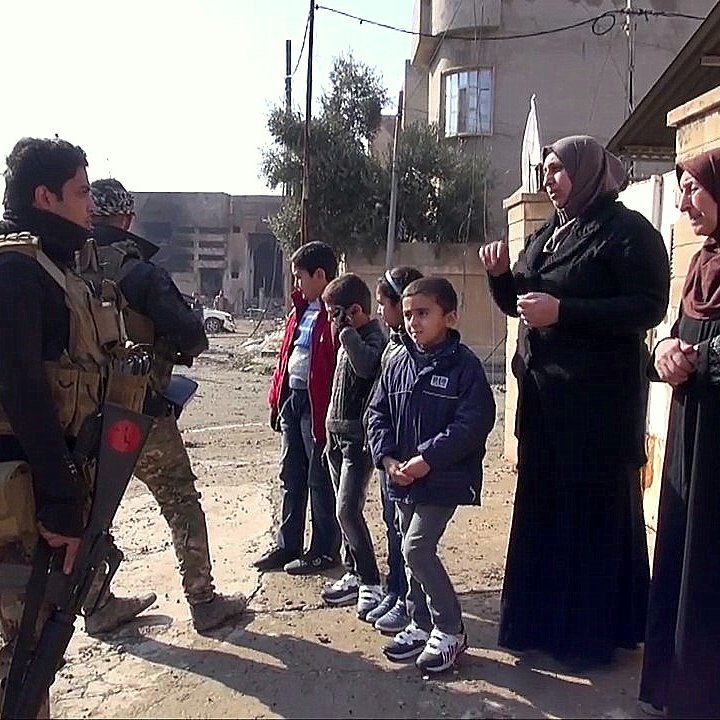 تظهر في الصورة مجموعة من الأشخاص في شارع يبدو أنه في منطقة تتعرض للتوتر أو النزاع. في الجهة اليسرى، يوجد جندي مسلح يرتدي زيًا عسكريًا، ويتحدث مع مجموعة من النساء والأطفال. النساء يرتدين ملابس تقليدية، بينما يبدو الأطفال مهتمين بالمحادثة. خلفهم، يمكن رؤية مباني متضررة وأجواء تعكس حالة المنطقة.