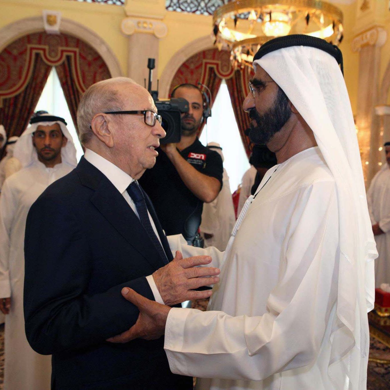 The image depicts two men engaging in conversation, likely in a formal setting. One man is dressed in a dark suit and glasses, while the other is in traditional Emirati attire, including a white kandura and headscarf. They appear to be having a cordial exchange, with the background showing a lavishly decorated room, including red curtains, ornate furniture, and a gathering of individuals in similar traditional attire. The atmosphere seems formal and respectful.