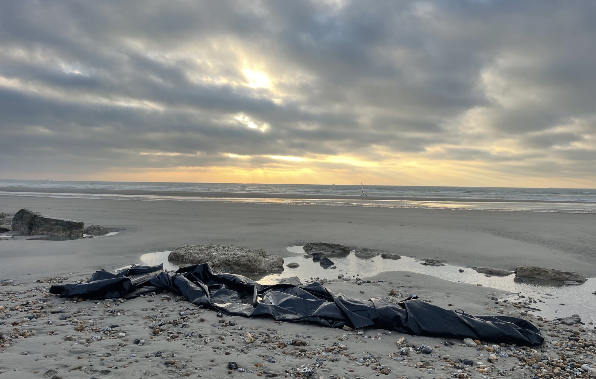 The image depicts a beach scene with a gray, cloudy sky creating a moody atmosphere. In the foreground, there is a large, black tarp or covering laid out on the sand, surrounded by smooth stones and scattered seashells. The beach appears to be wet, likely from recent tides, and there are gentle waves in the background. In the distance, a boat can be seen on the horizon, and rays of sunlight break through the clouds, creating a contrast with the darker tones of the sand and the tarp.