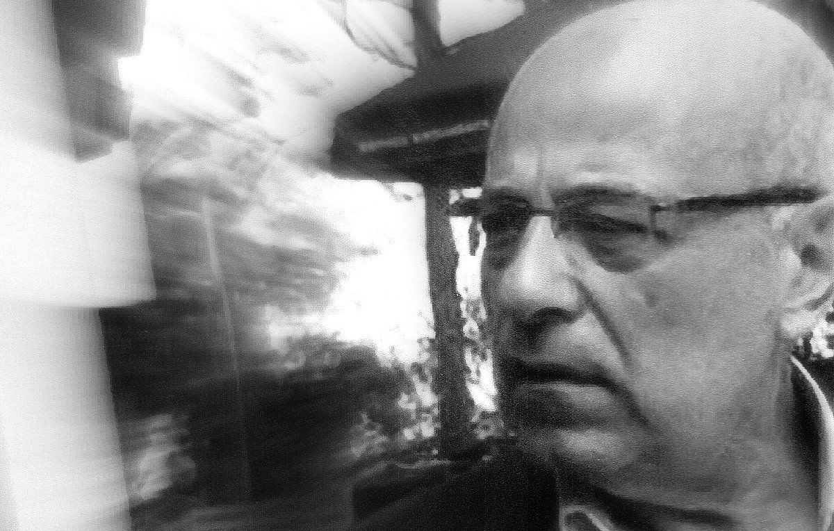 تظهر الصورة رجلًا في منتصف العمر، يبدو جادًا ومتأملًا. يعبر وجهه عن التفكير العميق أو الانشغال بشيء ما. يبدو أن الصورة قد أُخذت في مكان خارجي، حيث يمكن رؤية بعض العناصر الطبيعية أو المعمارية في الخلفية، مما يعطي إحساسًا بالعمق. الصورة تبدو مشوشة قليلاً، مما يضيف تأثيرًا فنيًا إليها.