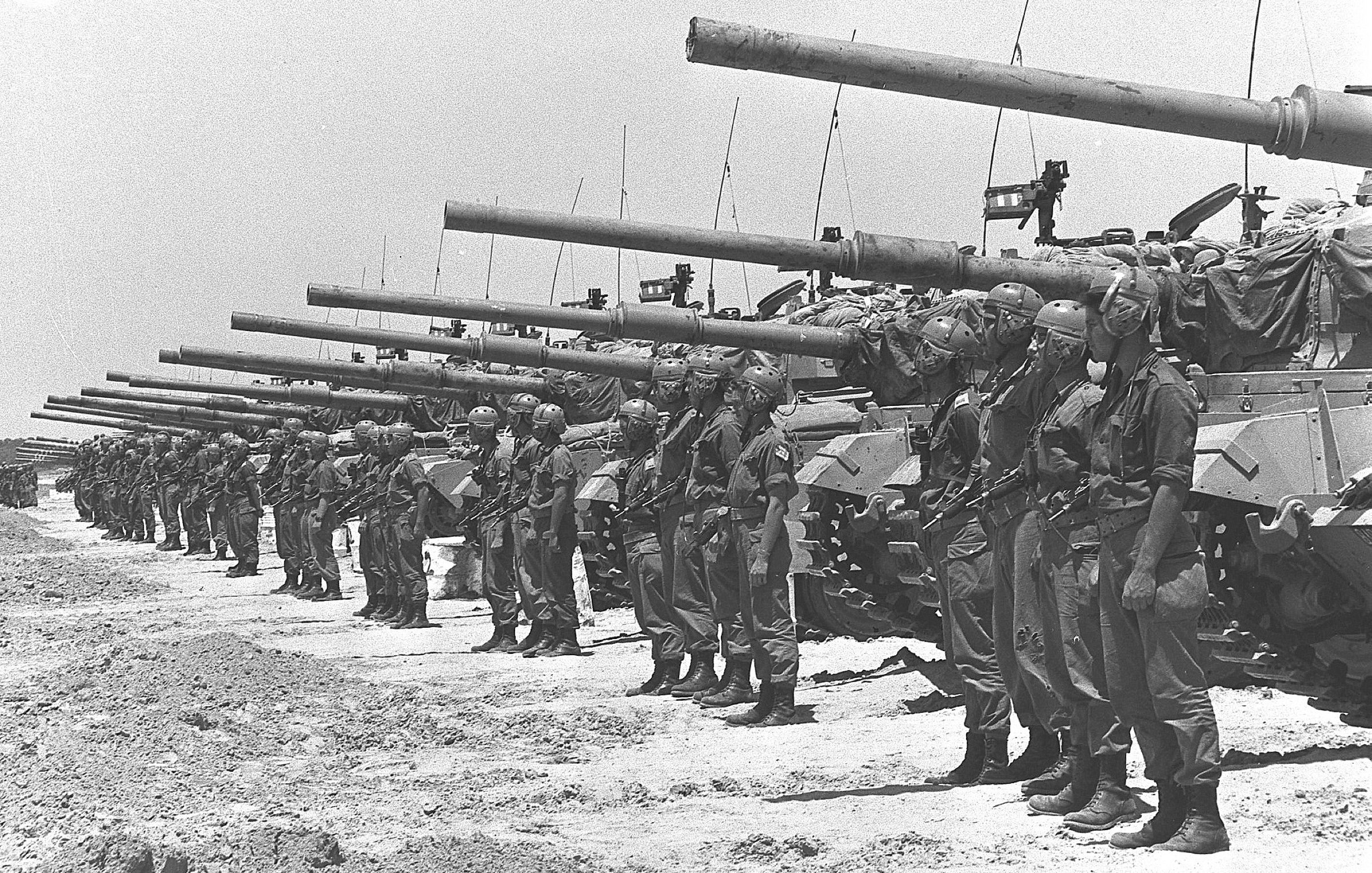 L'image montre un groupe de soldats alignés de manière formelle, portant des uniformes militaires et des casques. Ils sont positionnés devant une rangée de chars d'assaut, qui sont également alignés. Le paysage semble désertique, sous un ciel clair. L'atmosphère évoque une préparation militaire, peut-être pour une parade ou une démonstration de force. Les chars sont équipés de canons pointés vers l'avant, ce qui ajoute à l'impression de puissance et de militarisation de la scène.