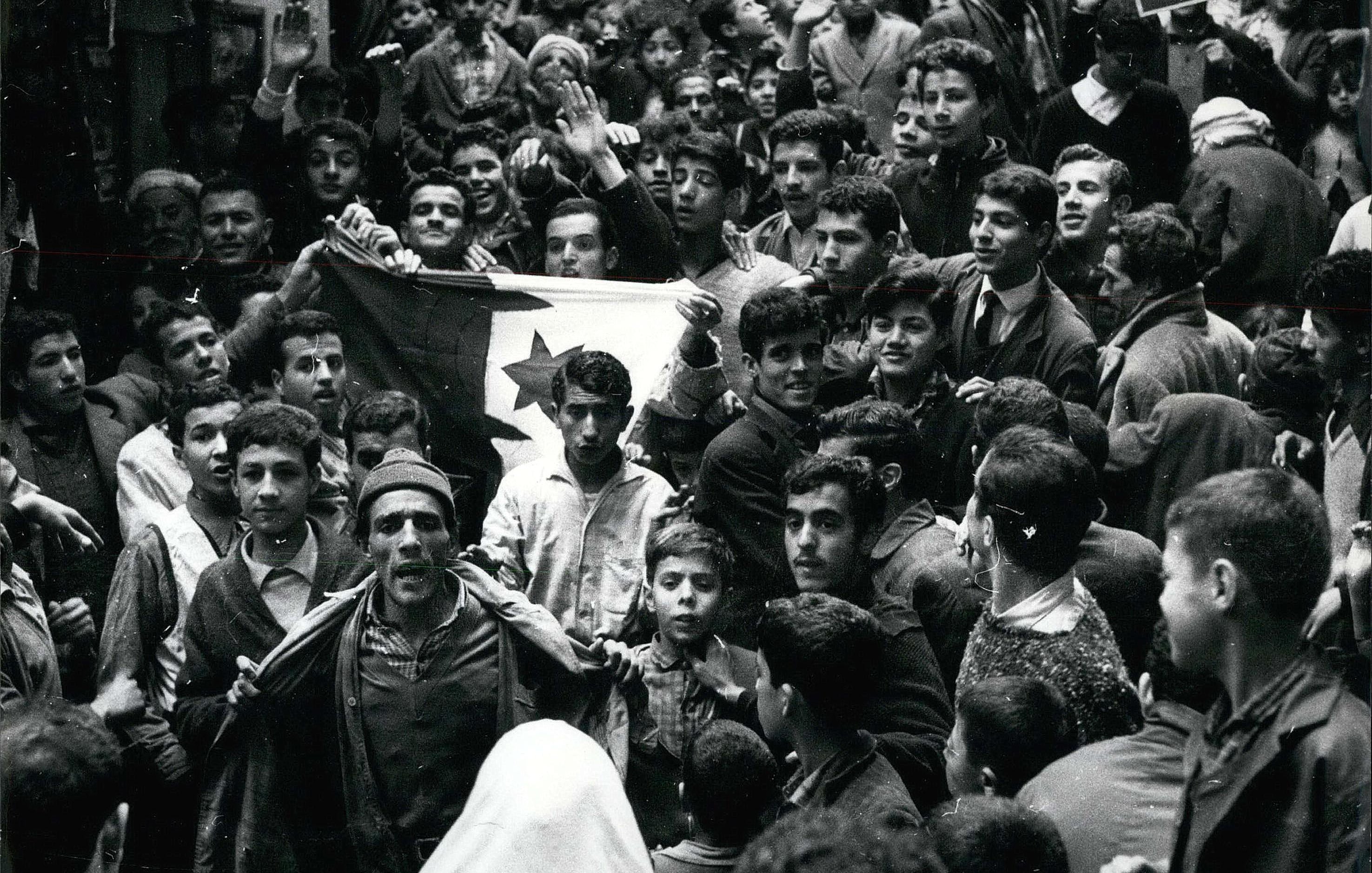 L'image montre une foule dense de personnes rassemblées dans une rue. Au premier plan, plusieurs individus sont visibles, dont un homme qui semble porter un drapeau avec un symbole distinctif. Les expressions des visages varient, certains levant les bras en signe de soutien ou d'enthousiasme. On peut observer des jeunes, des adultes et même des enfants parmi la foule. L'atmosphère semble intense et chargée d'émotion, indiquant un moment significatif ou une manifestation. La scène est en noir et blanc, ce qui lui donne un aspect historique.