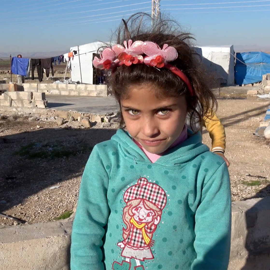 تظهر في الصورة فتاة صغيرة ترتدي سترة زرقاء مزينة بصورة لفتاة أخرى. لديها عيون فاتحة ومشرقة وشعر داكن، مع زينة زهرية في شعرها. الخلفية تضم خيام وغير منظمة، مما يوحي بأنها في مخيم. تبدو الفتاة مبتسمة ومرحة.